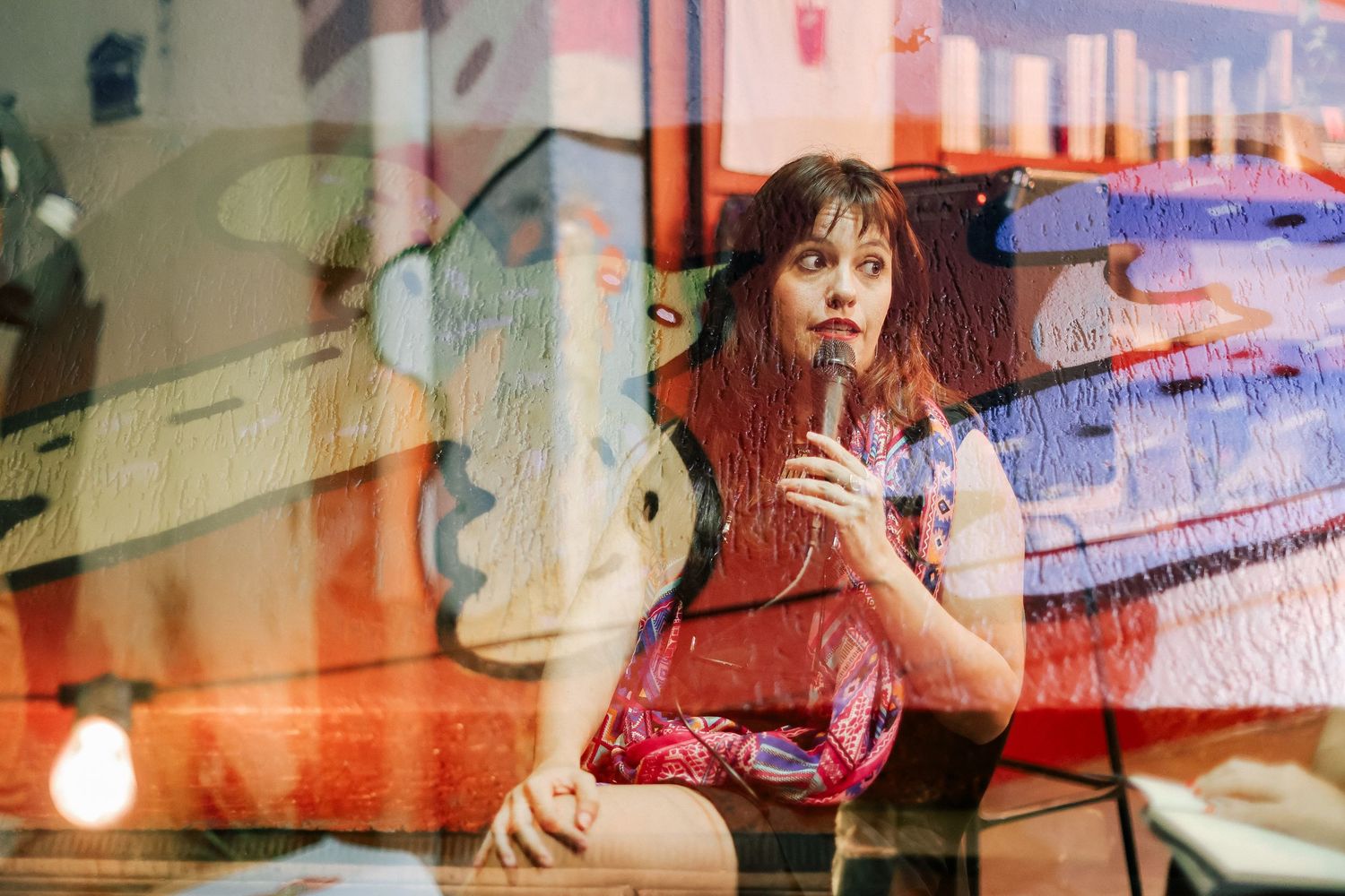 Young woman with red hair speaks into a microphone seated against a colorful abstract mural backdrop indoors.