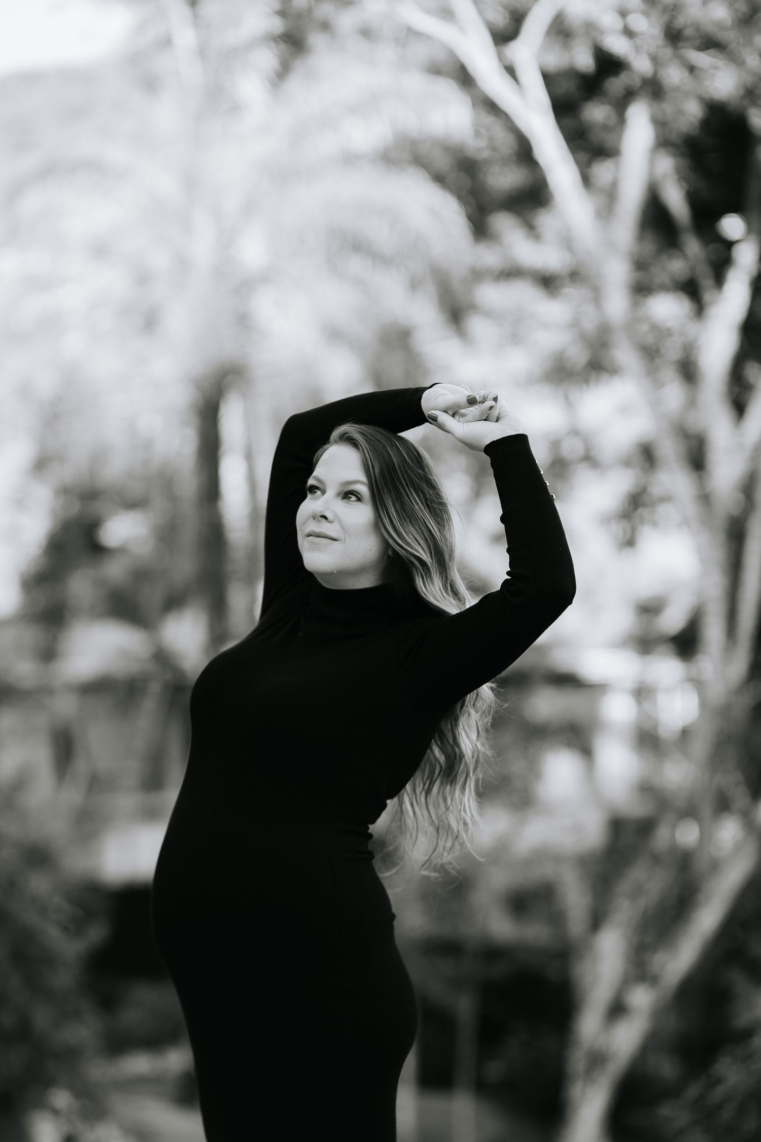 Dramatic black and white outdoor maternity photo with woman in black dress posing against trees.