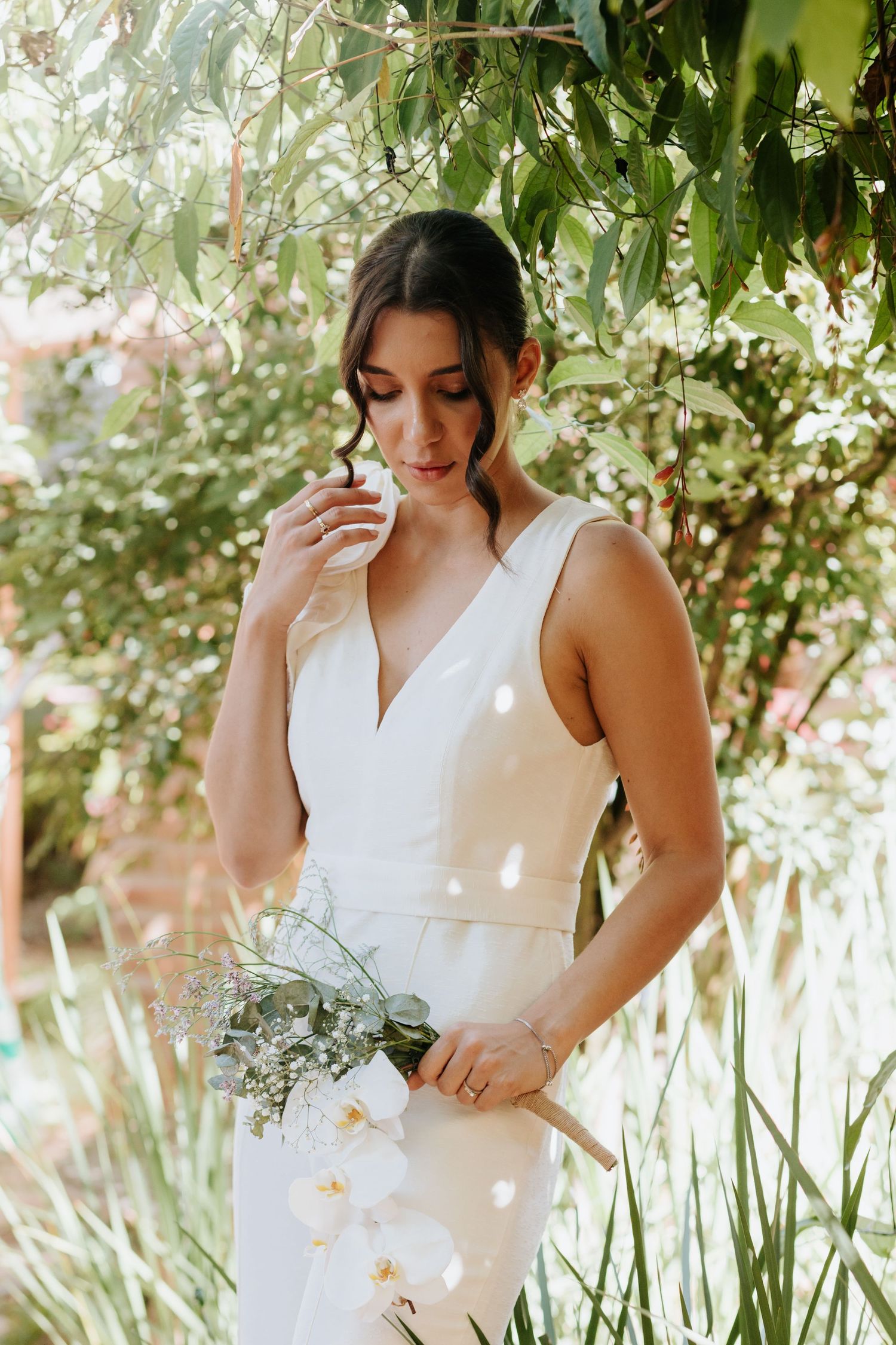 A person in a sleeveless white dress holding a delicate bouquet in a sunlit garden setting.