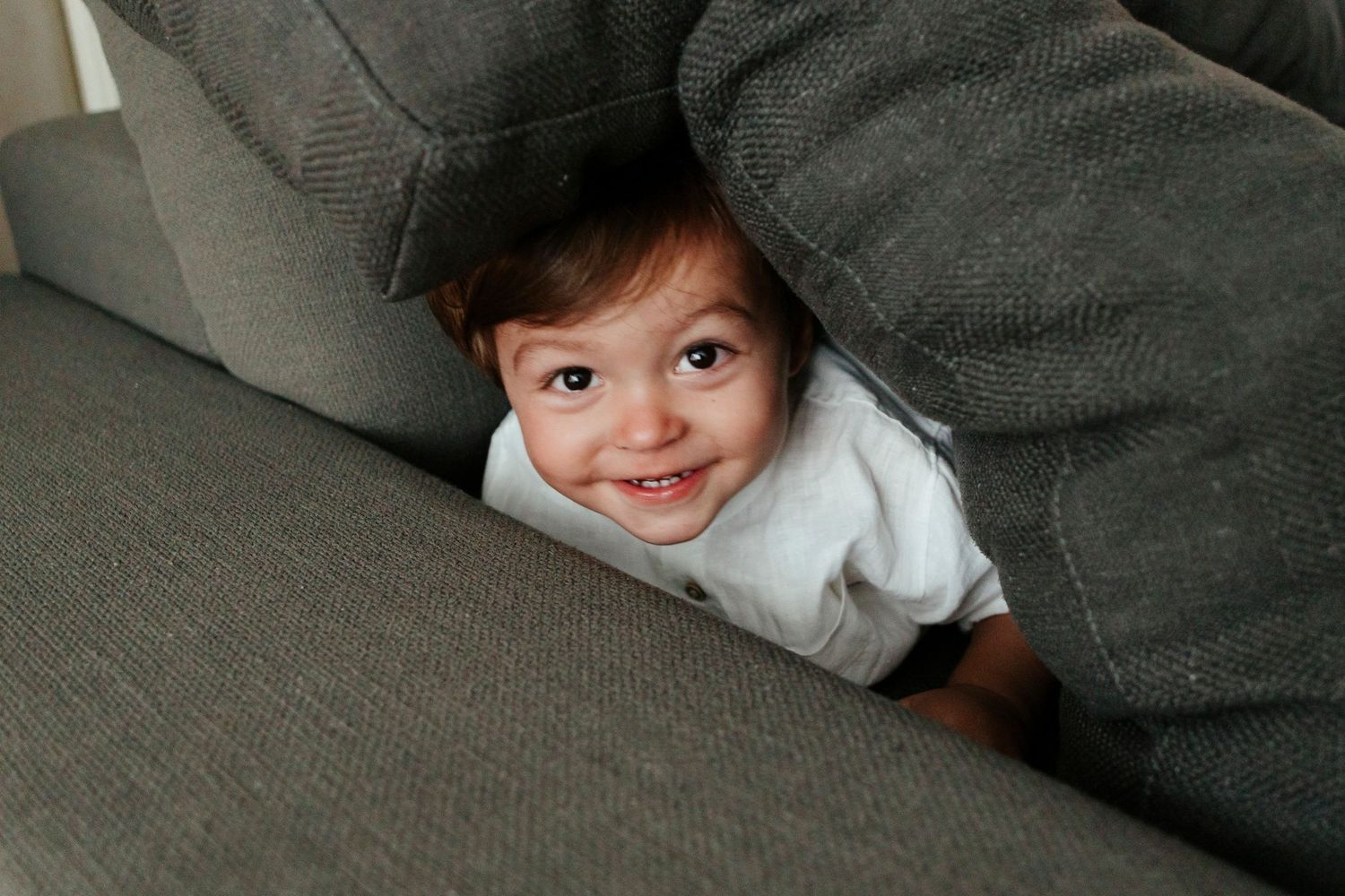 Playful toddler peeks out from between couch cushions with a bright smile.