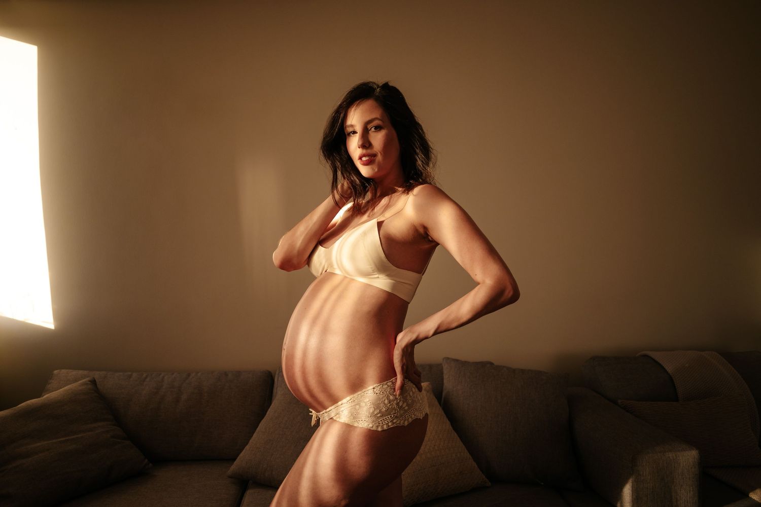 Dramatic side pose maternity photo with strong window lighting and cream colored lingerie.