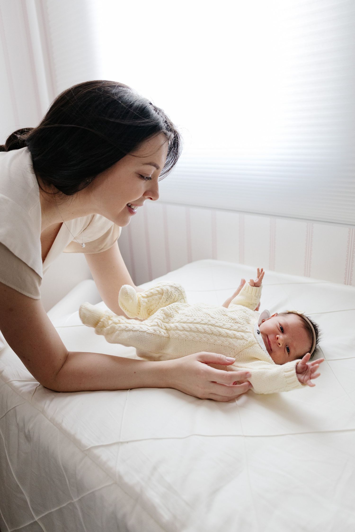 Mother leaning over crib smiling at newborn baby in cream knit outfit.