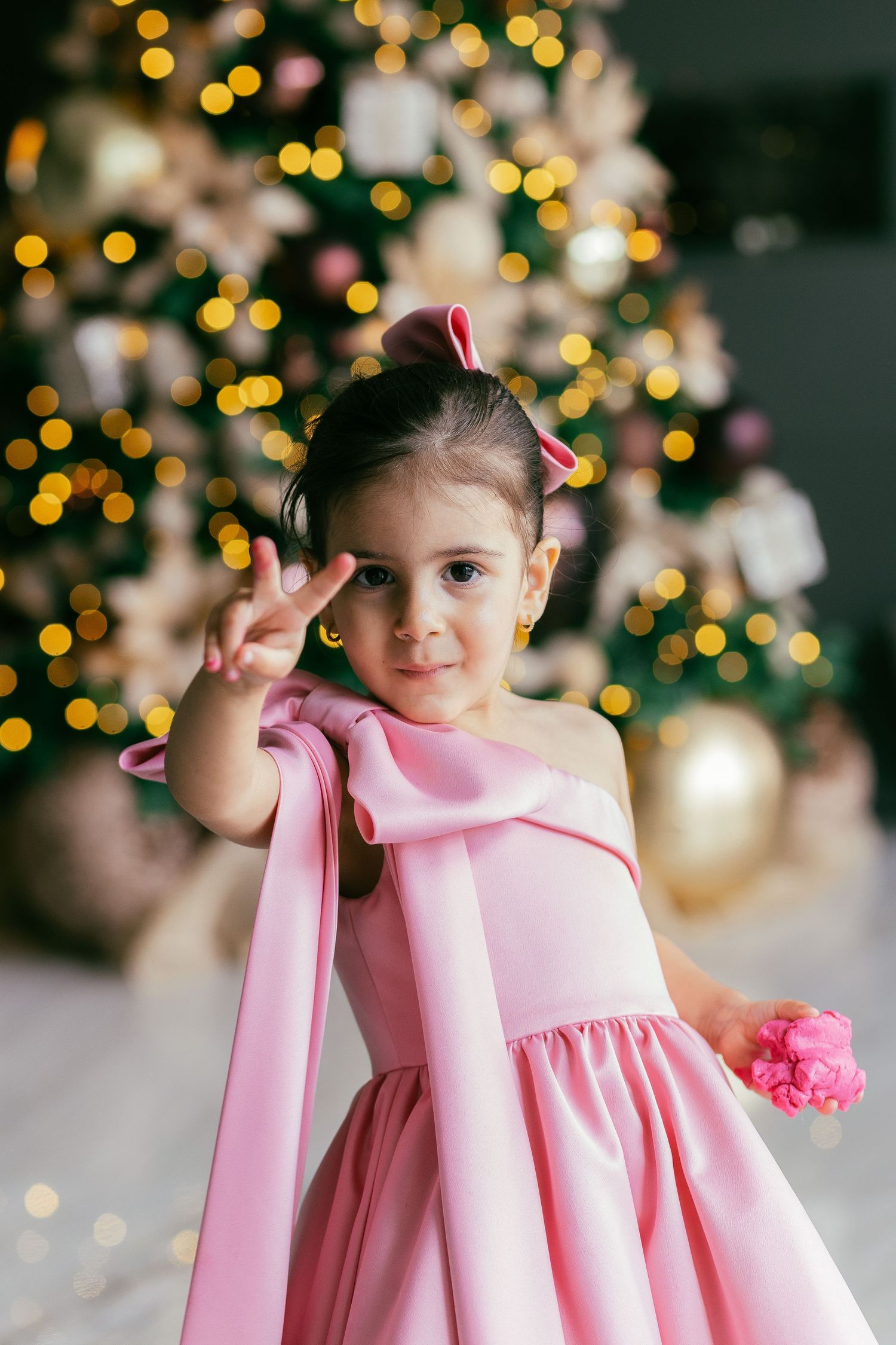 A child in a pink dress poses in front of a Christmas tree with twinkling bokeh lights in the background.