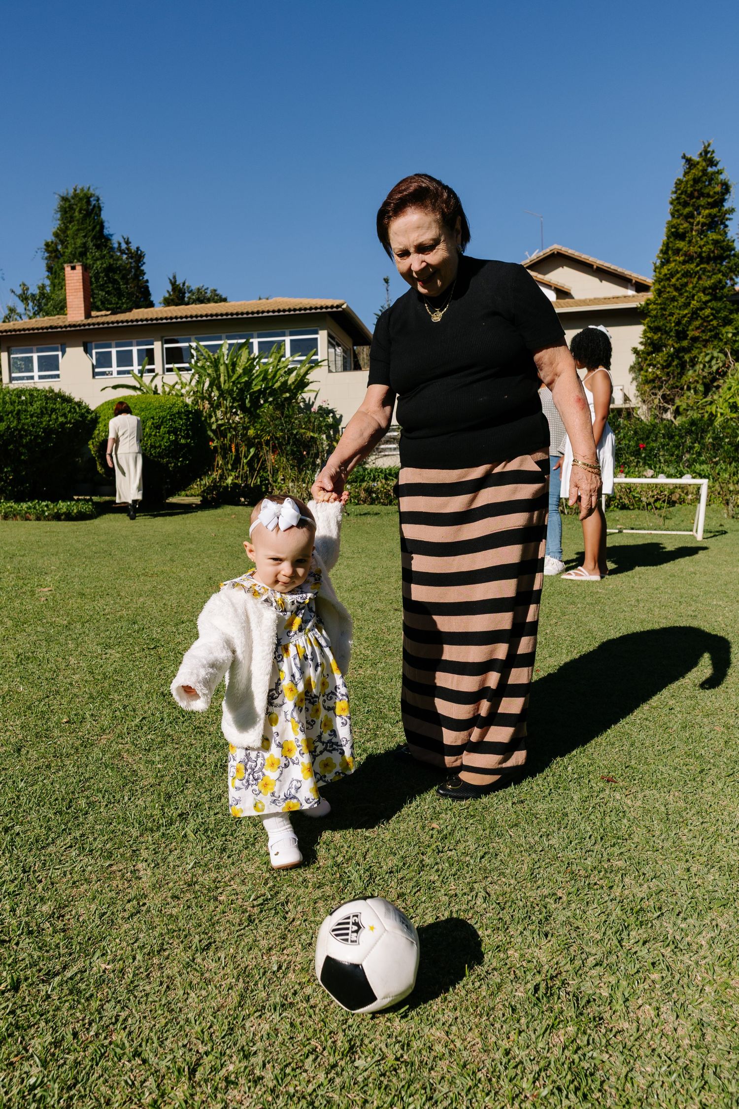 A parent and toddler playing soccer together on a sunny day in front of a suburban house.
