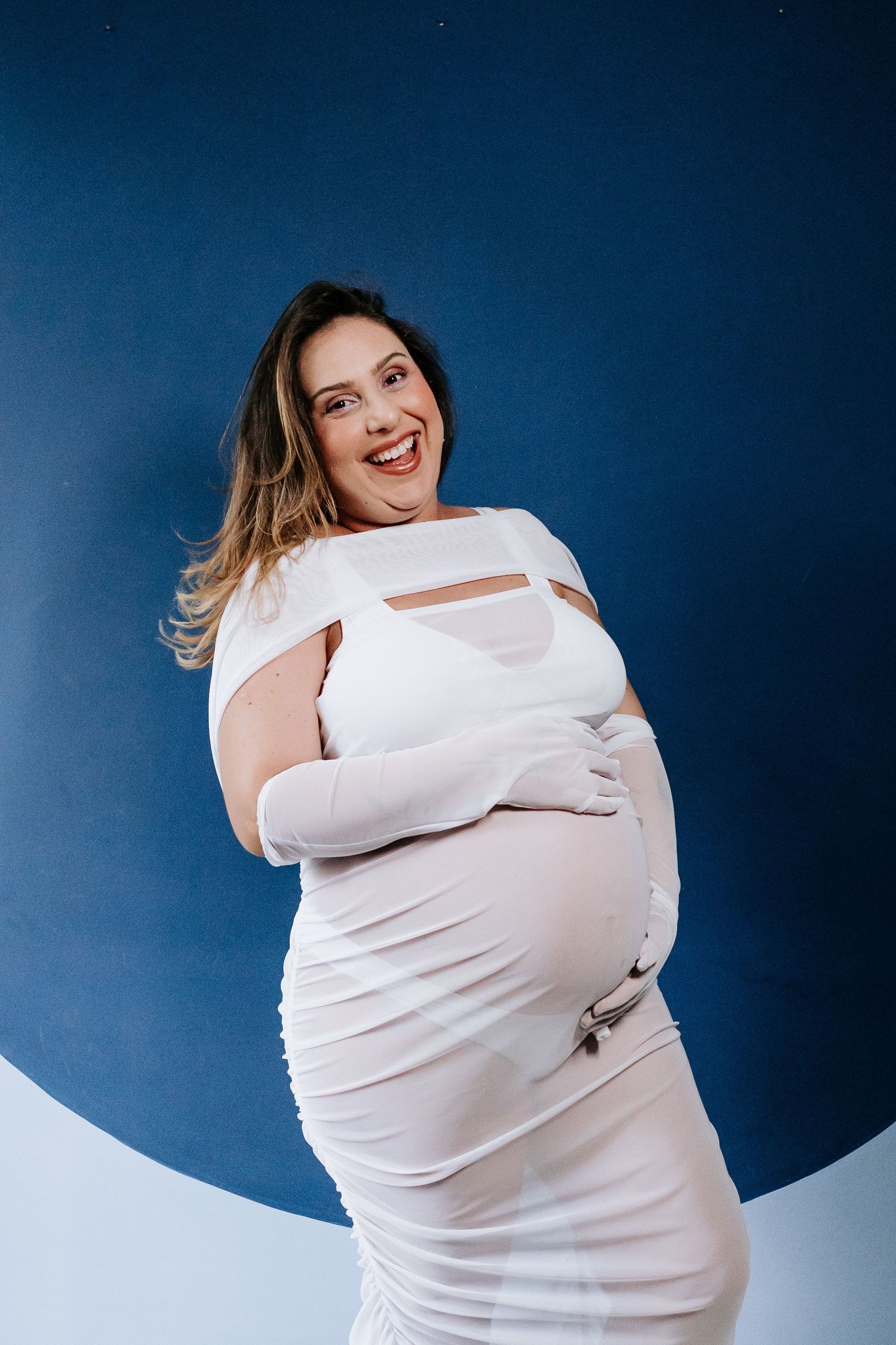 A joyful maternity portrait in white dress against a dramatic blue and white backdrop.
