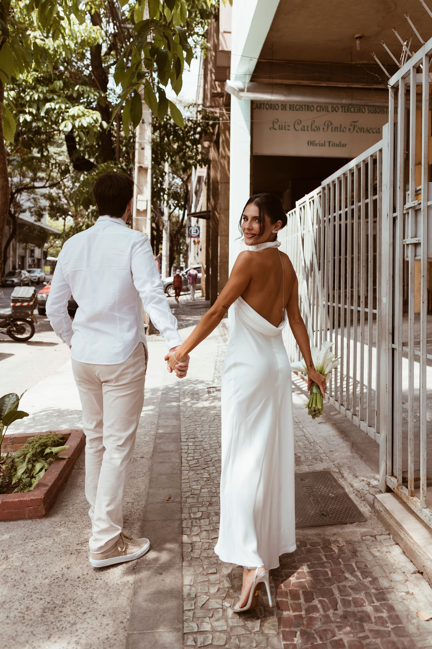 Couple in white outfits hold hands while walking down city sidewalk.