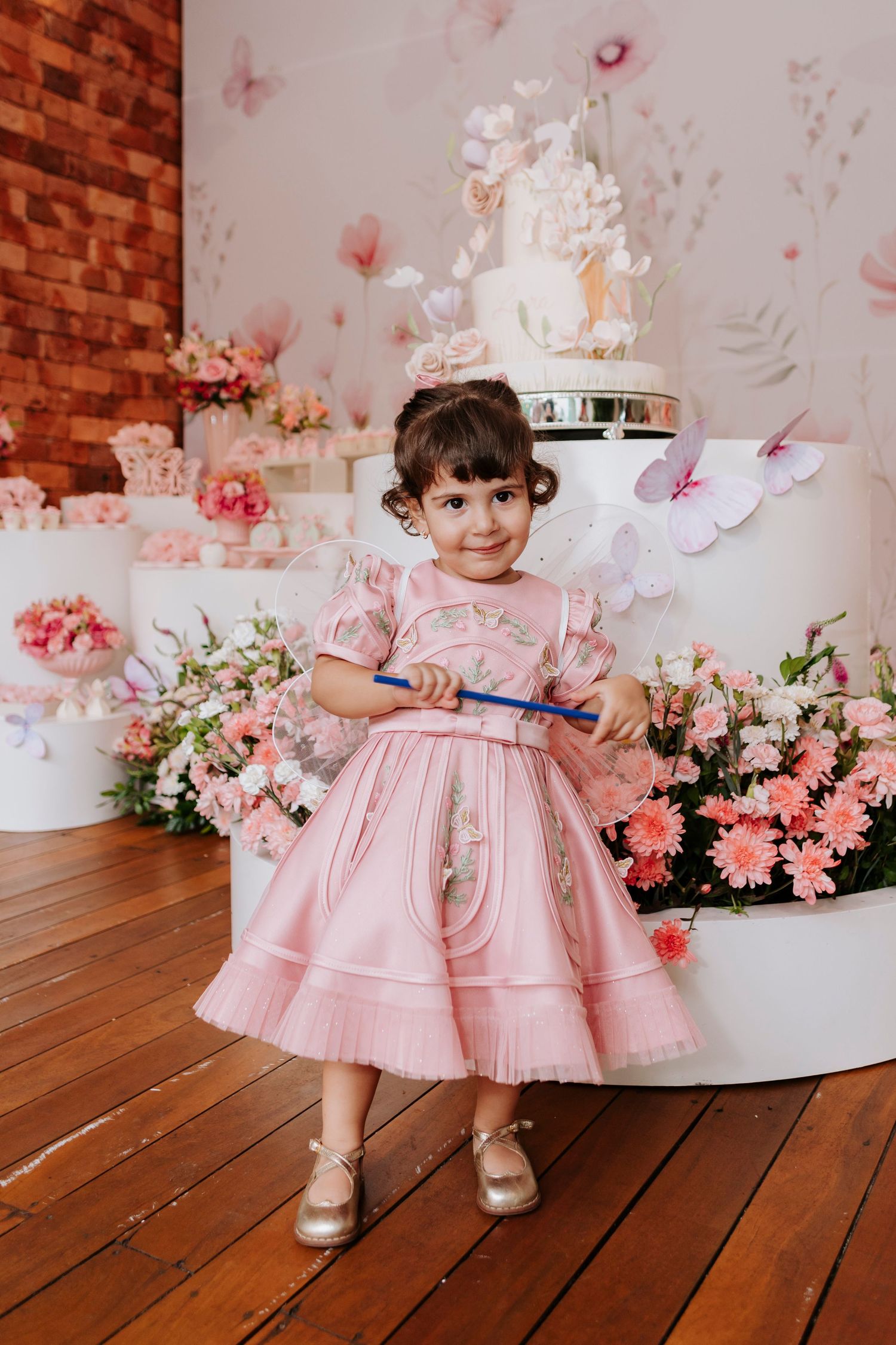 Child in pink ruffled dress posing against romantic backdrop with flowers and butterflies.