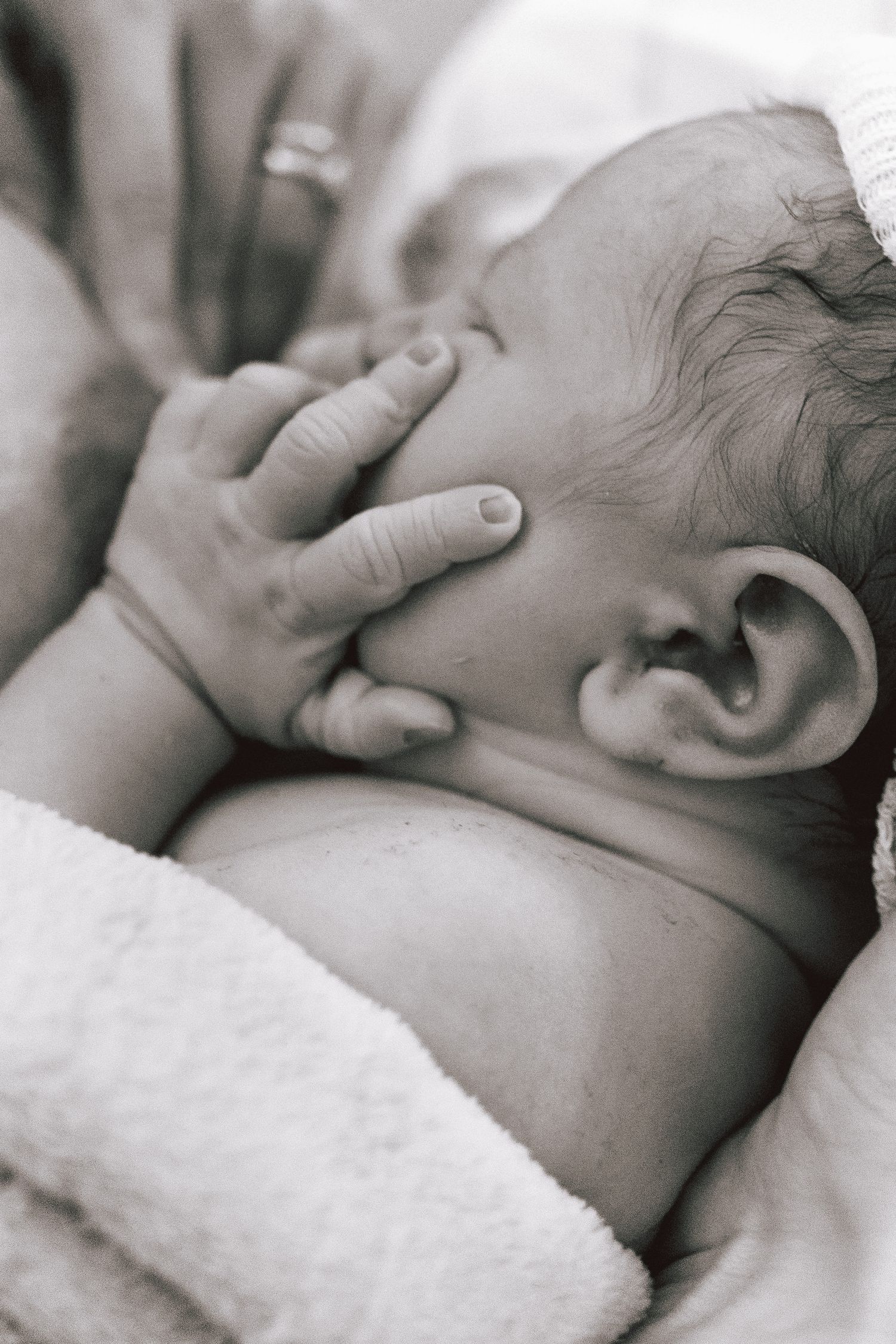 Black and white photo of a newborn baby cradled in an adult's arms, gently touching their own face.