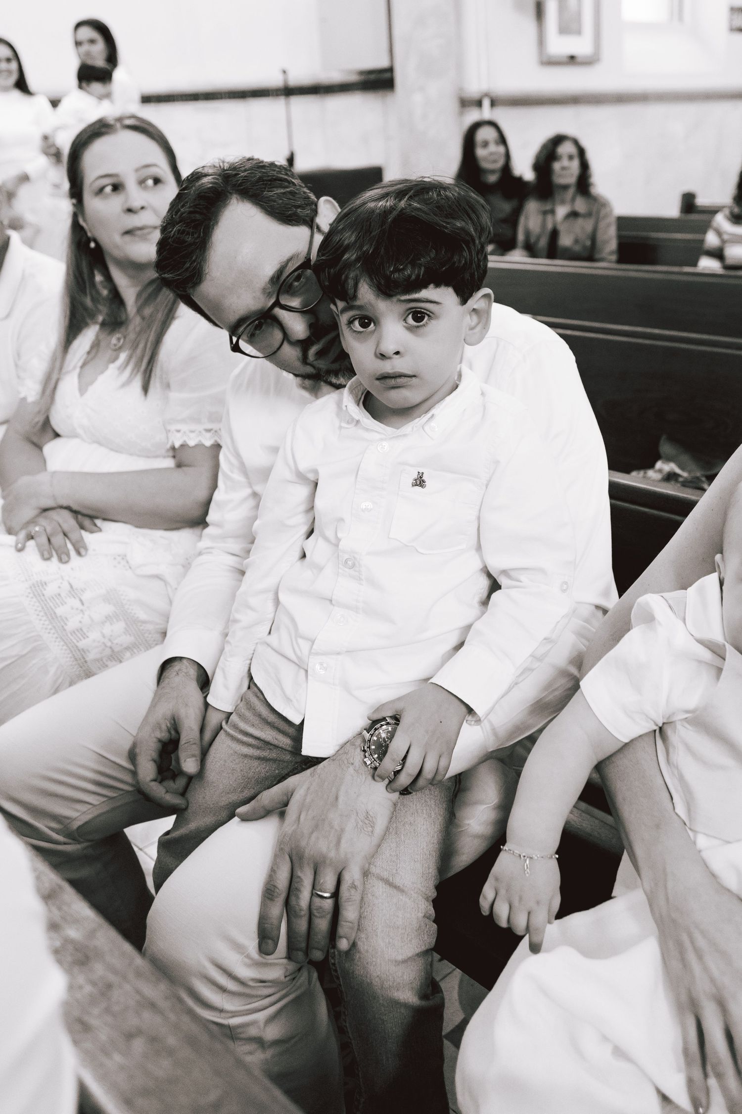 Children in white formal attire sit together on wooden church pews during a ceremony.