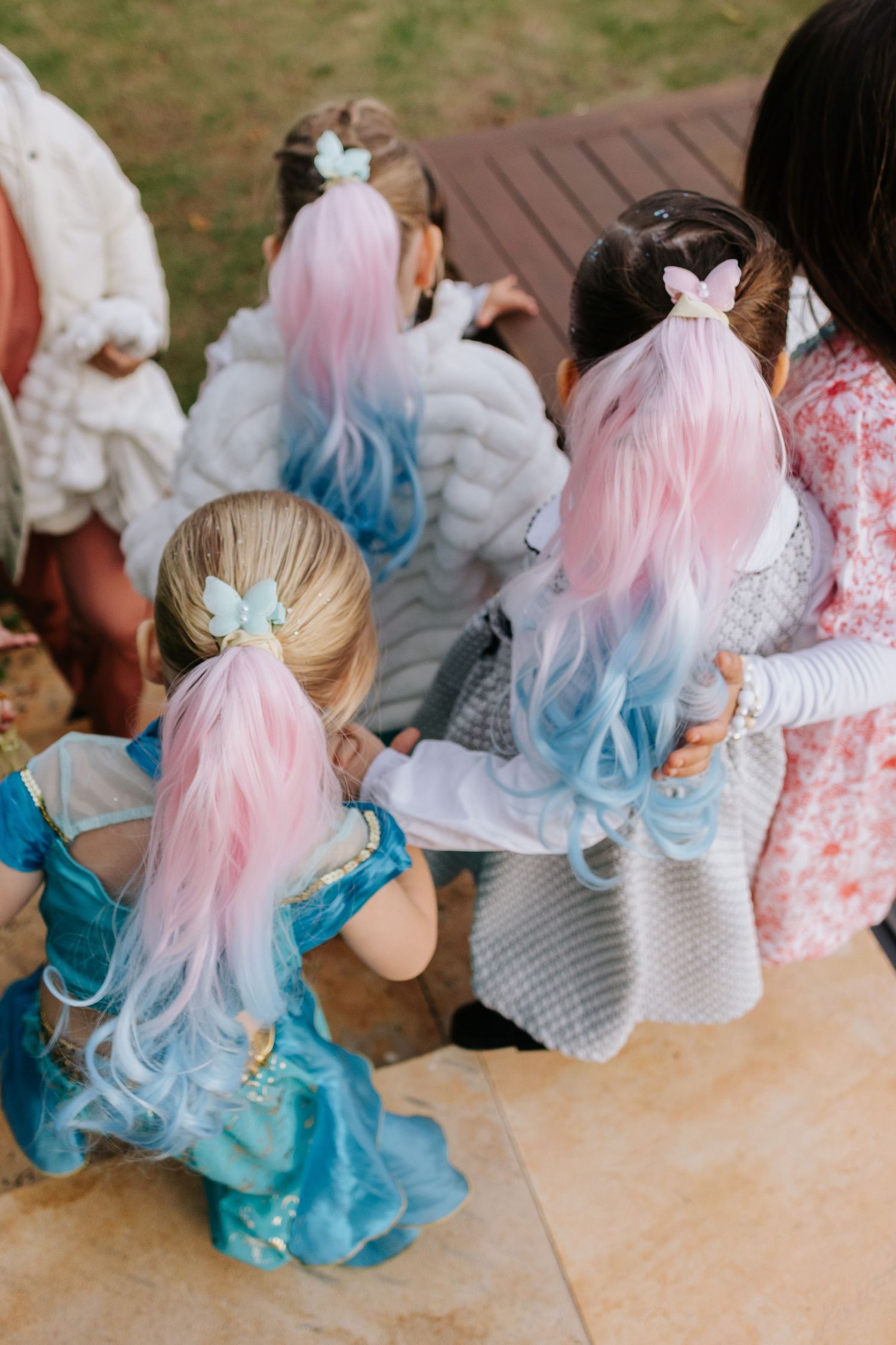 Children wearing matching pink and blue ombre hair extensions sit together at an outdoor party or celebration.