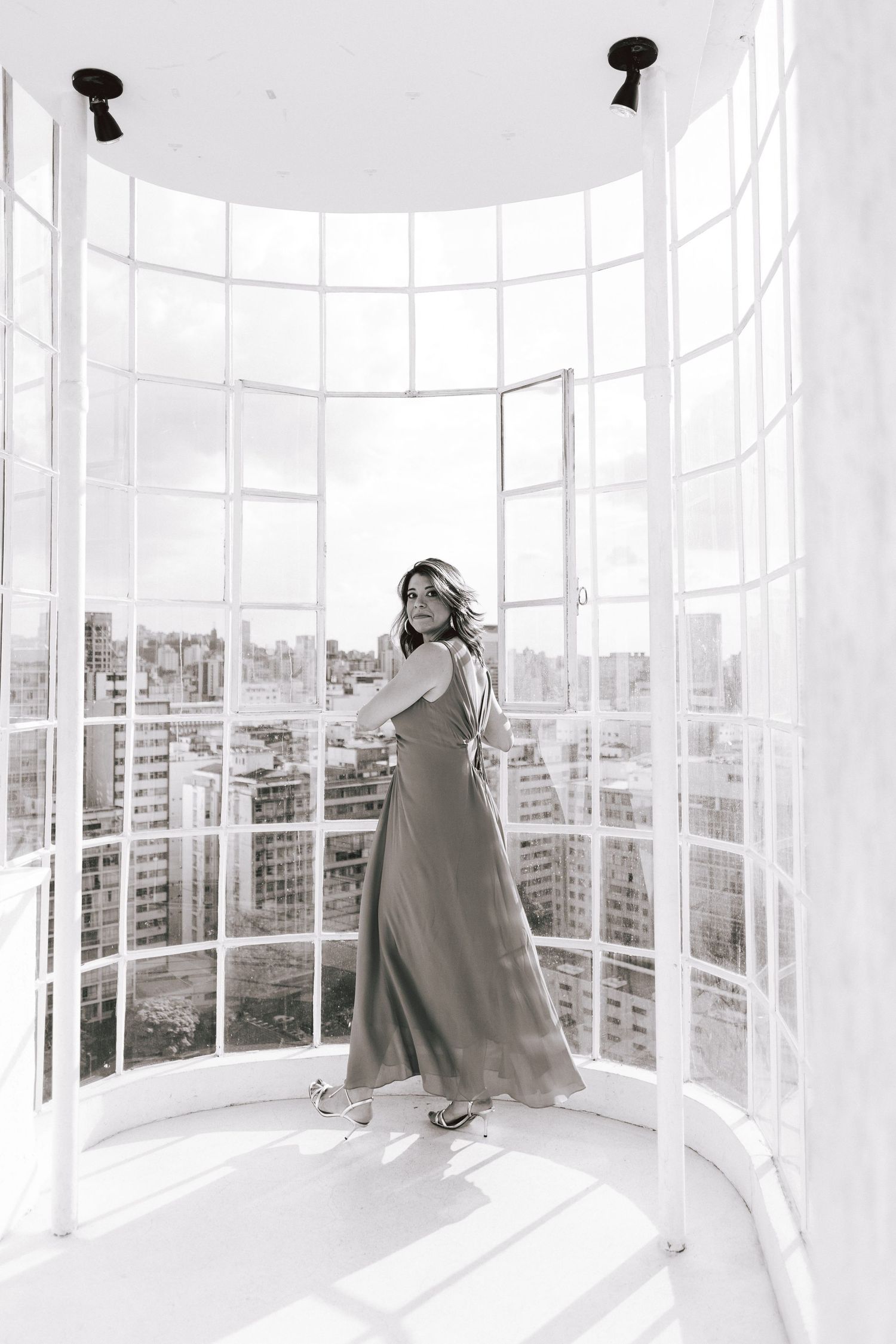 A curved glass-walled room with panoramic city views shows a figure in a long coat looking out at the urban landscape.
