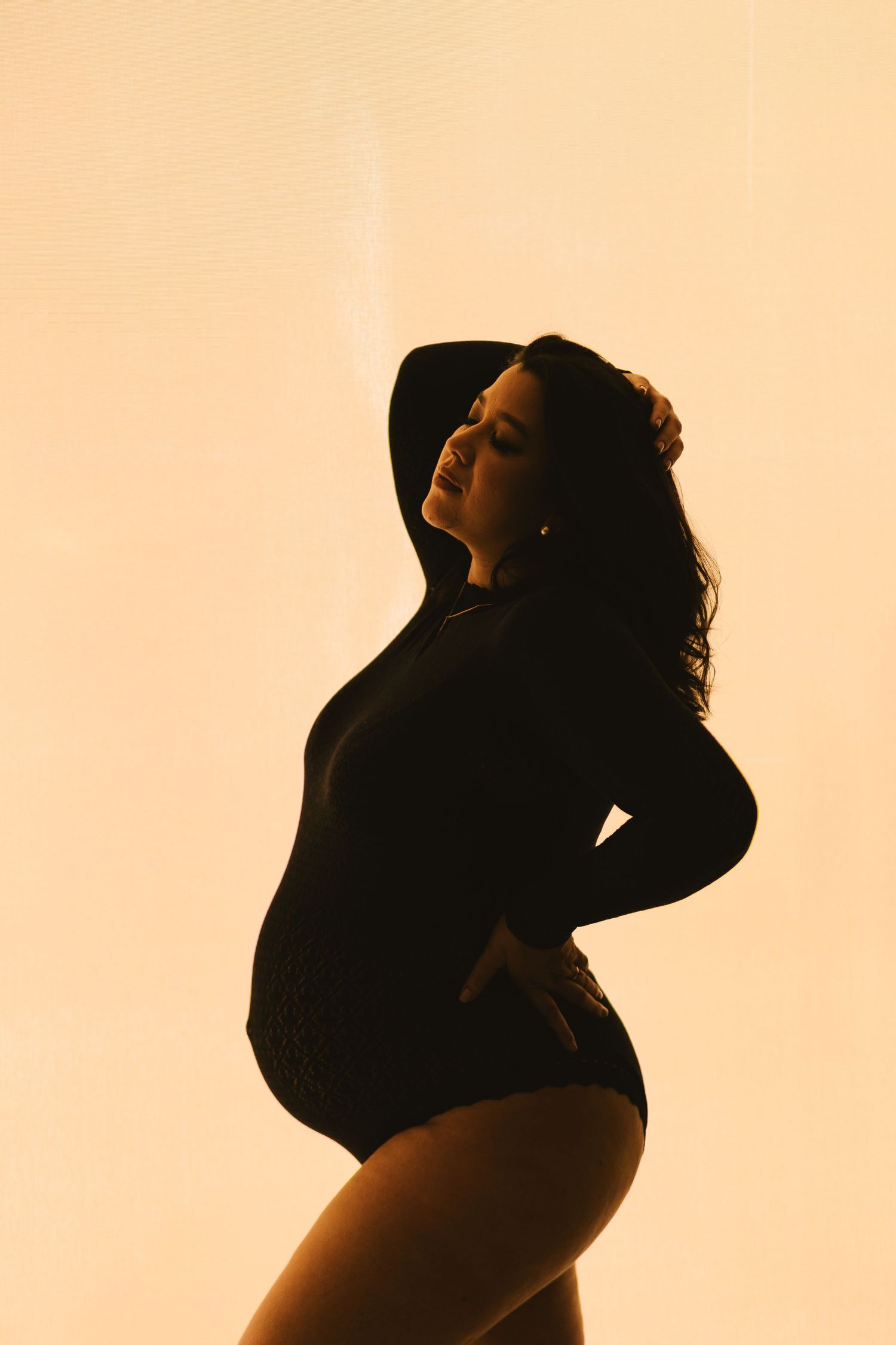 Silhouette of a pregnant woman in a black bodysuit posing gracefully against a warm golden backlit background.