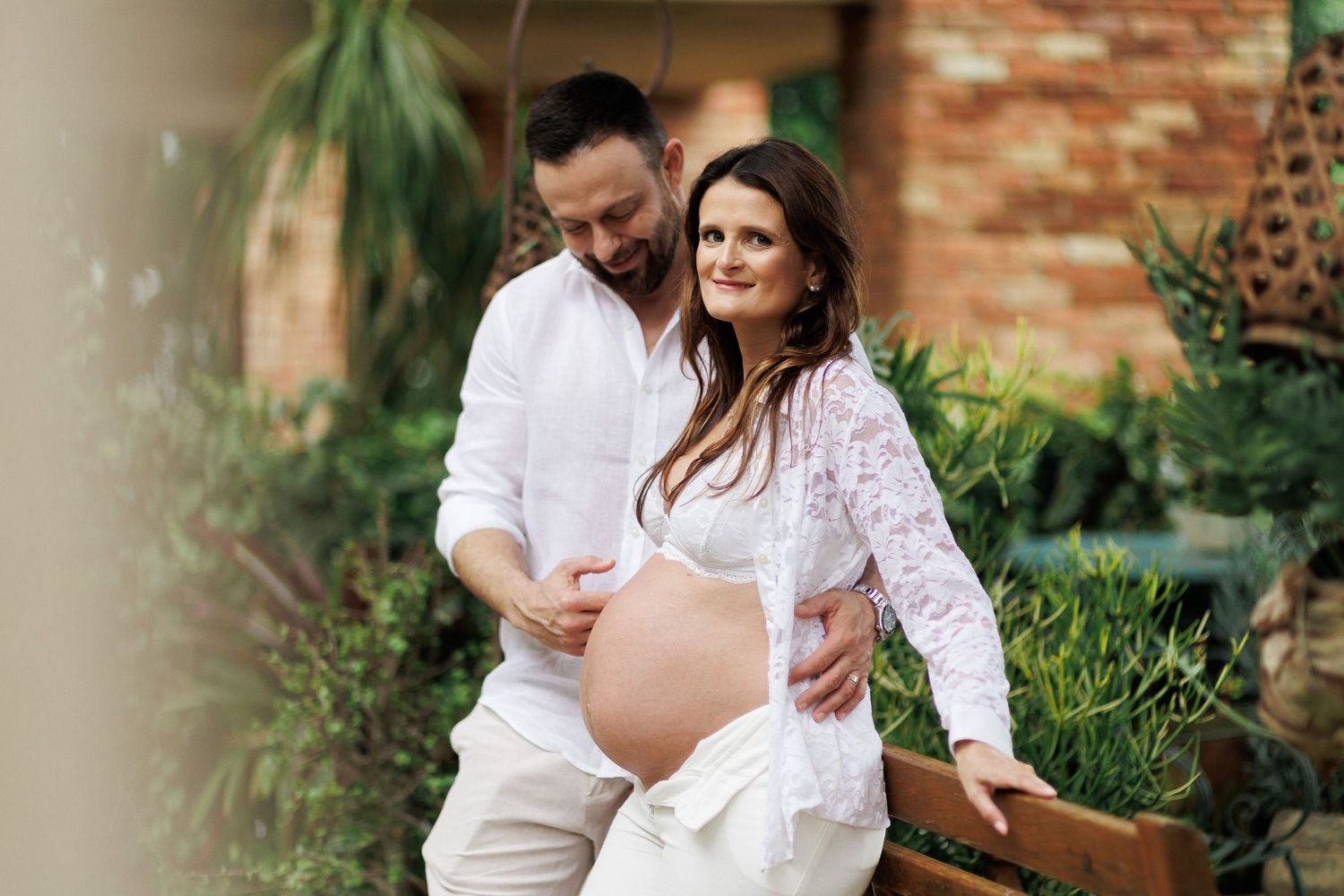 Couple posing for maternity photo shoot outdoors, man in white shirt embracing pregnant woman in lace top.