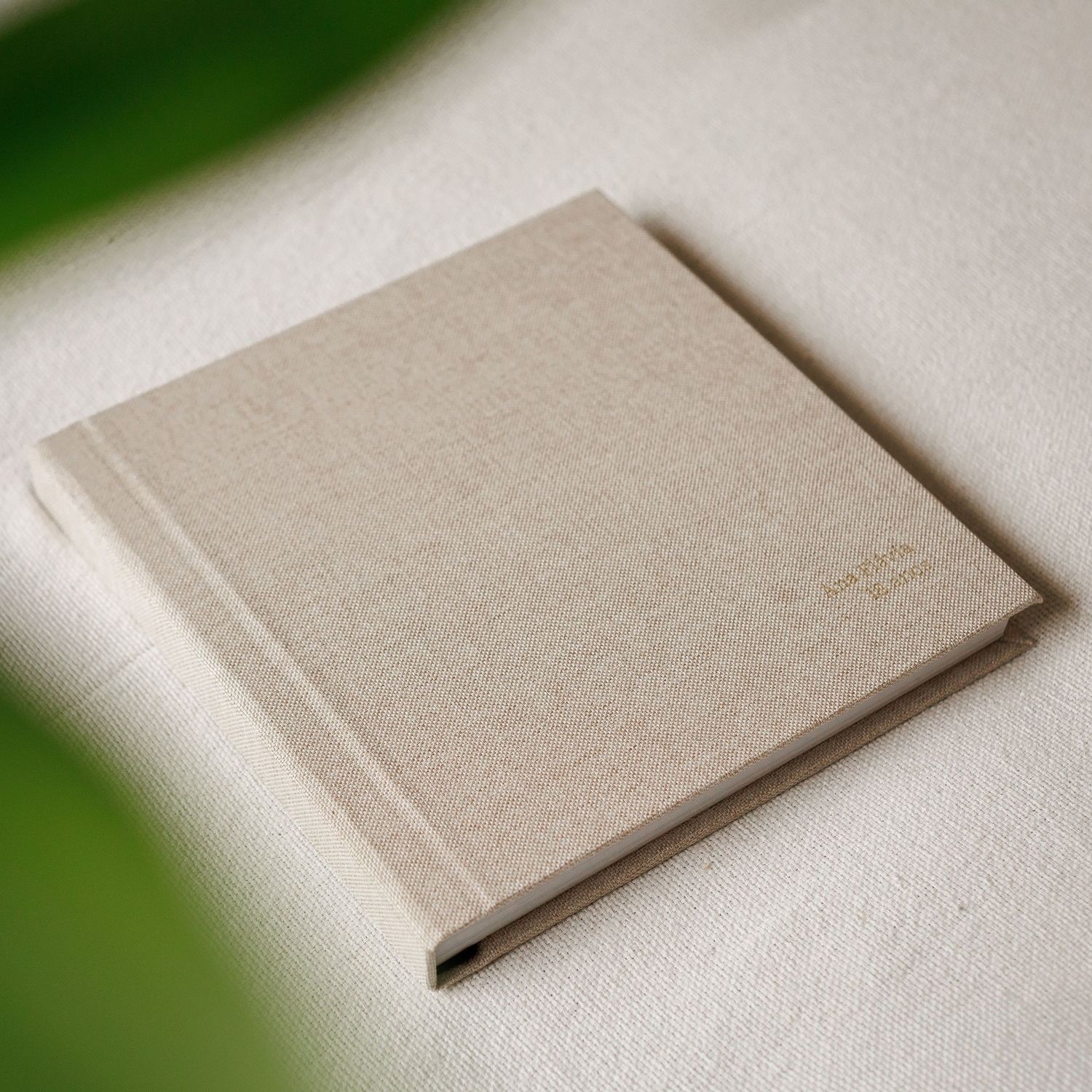 A square linen-covered hardcover photo album in natural beige with subtle gold embossing, resting on a white fabric surface.