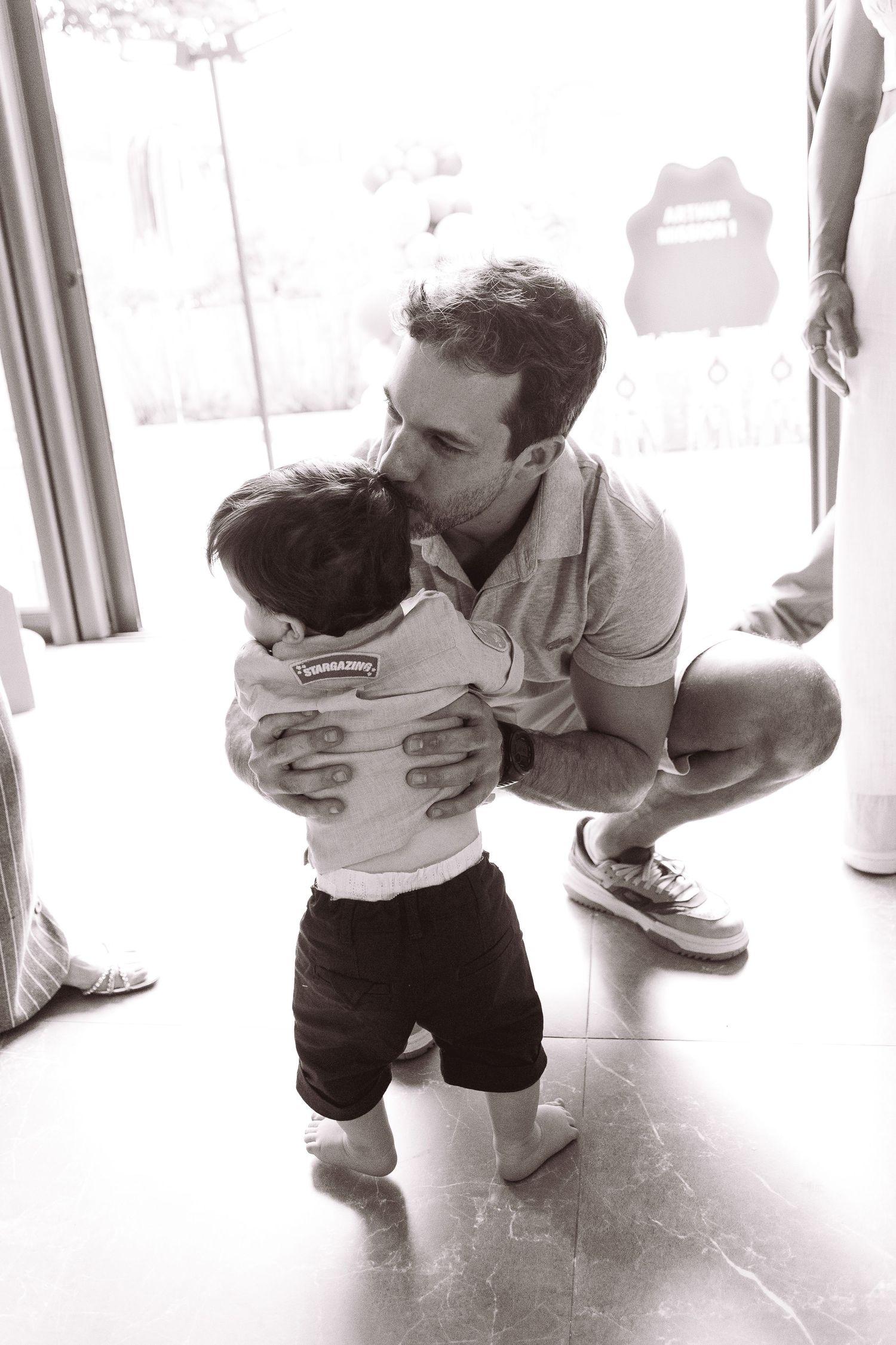 Black and white photo of a father kissing his toddler child while crouching down indoors.