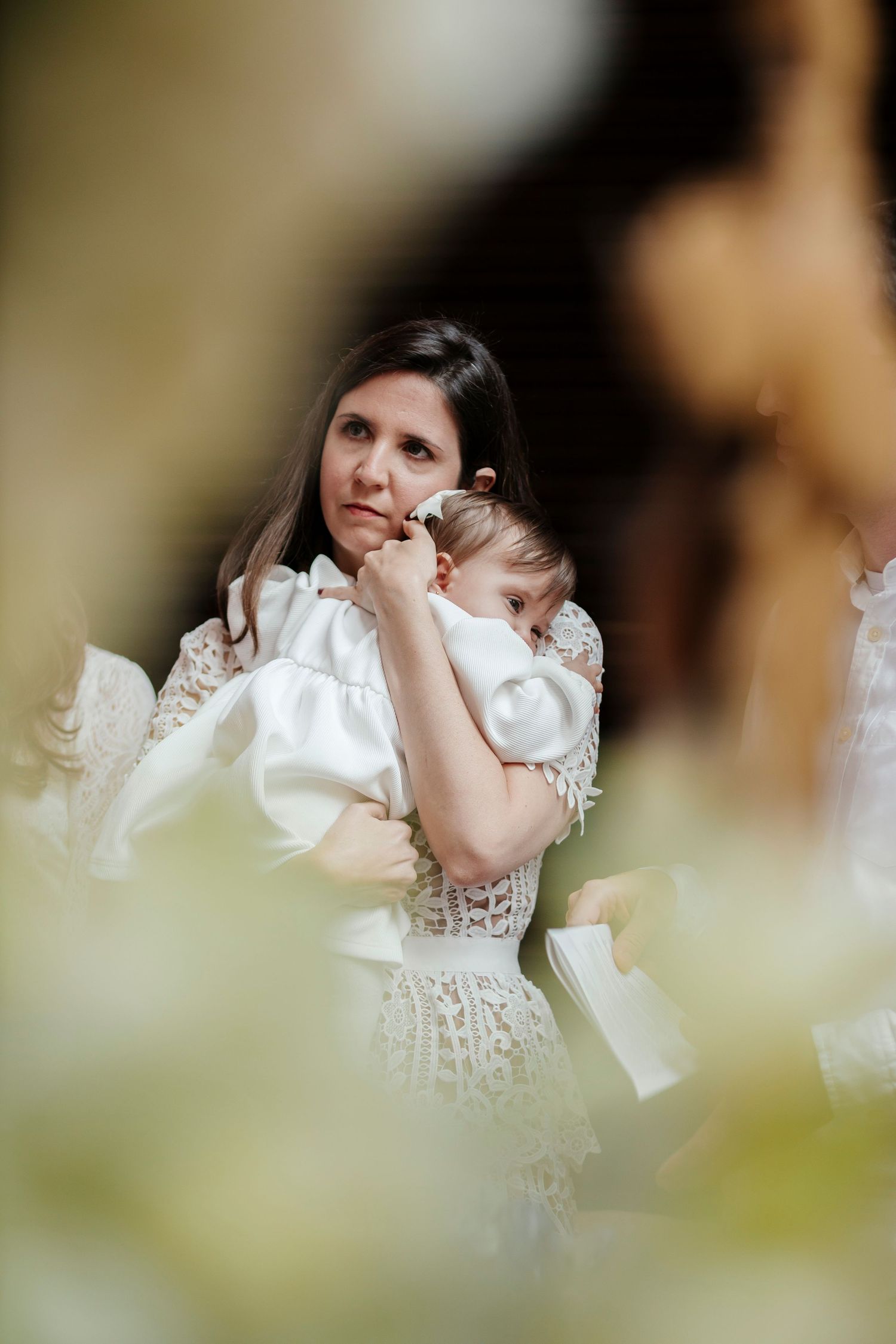 Woman in white lace dress holding sleeping baby during formal event.