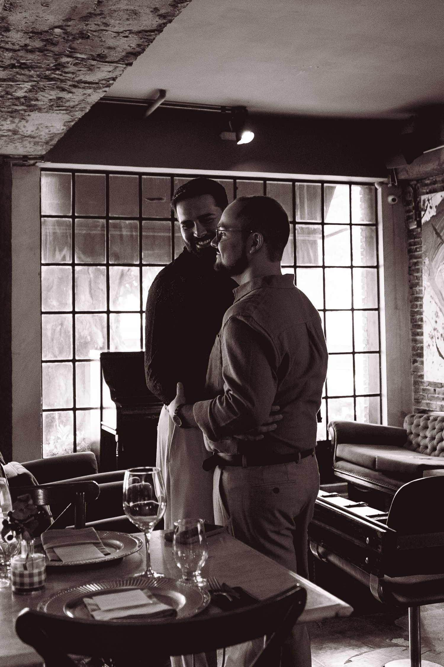 Two men share an intimate moment in a moody black and white restaurant setting with large industrial windows.
