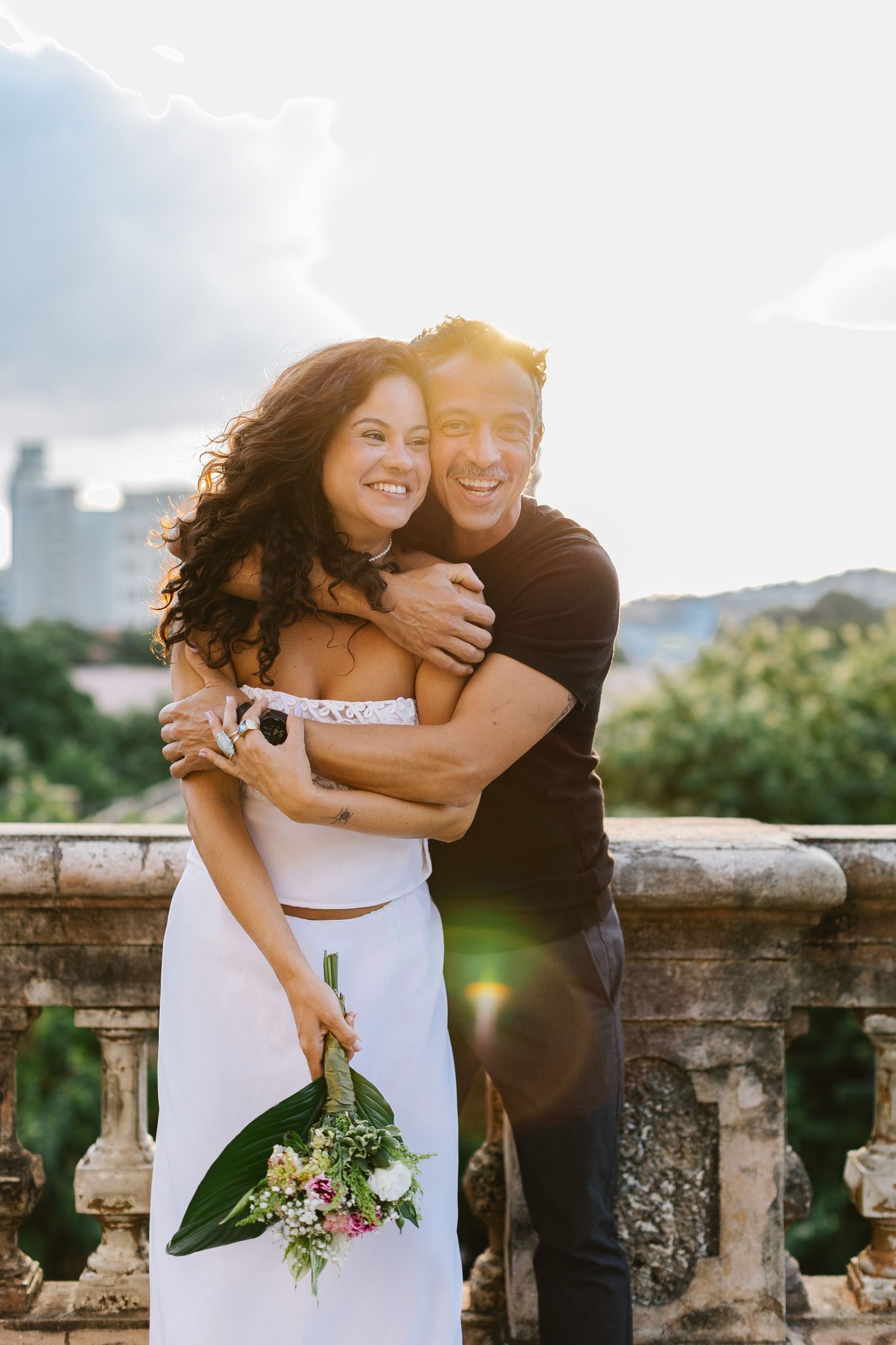 Smiling couple embracing on a stone balcony at sunset, woman holding a bouquet of flowers.