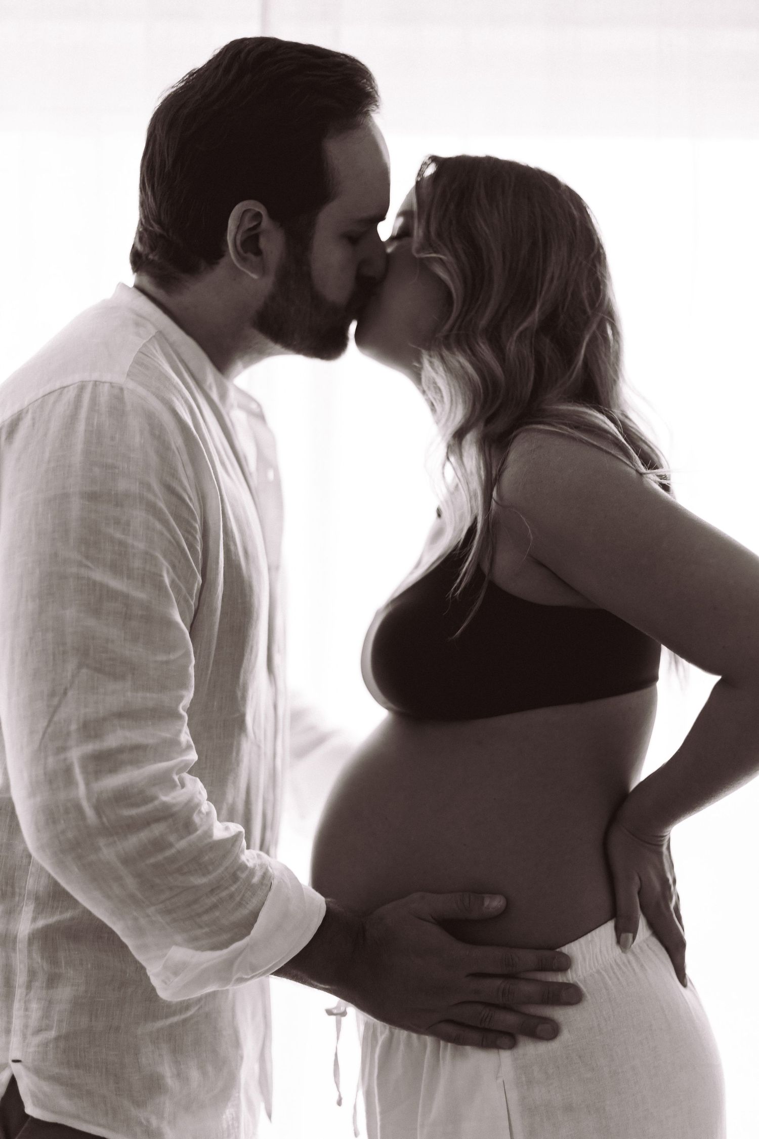 Black and white photo of a couple kissing while cradling a pregnant belly during a maternity photoshoot.