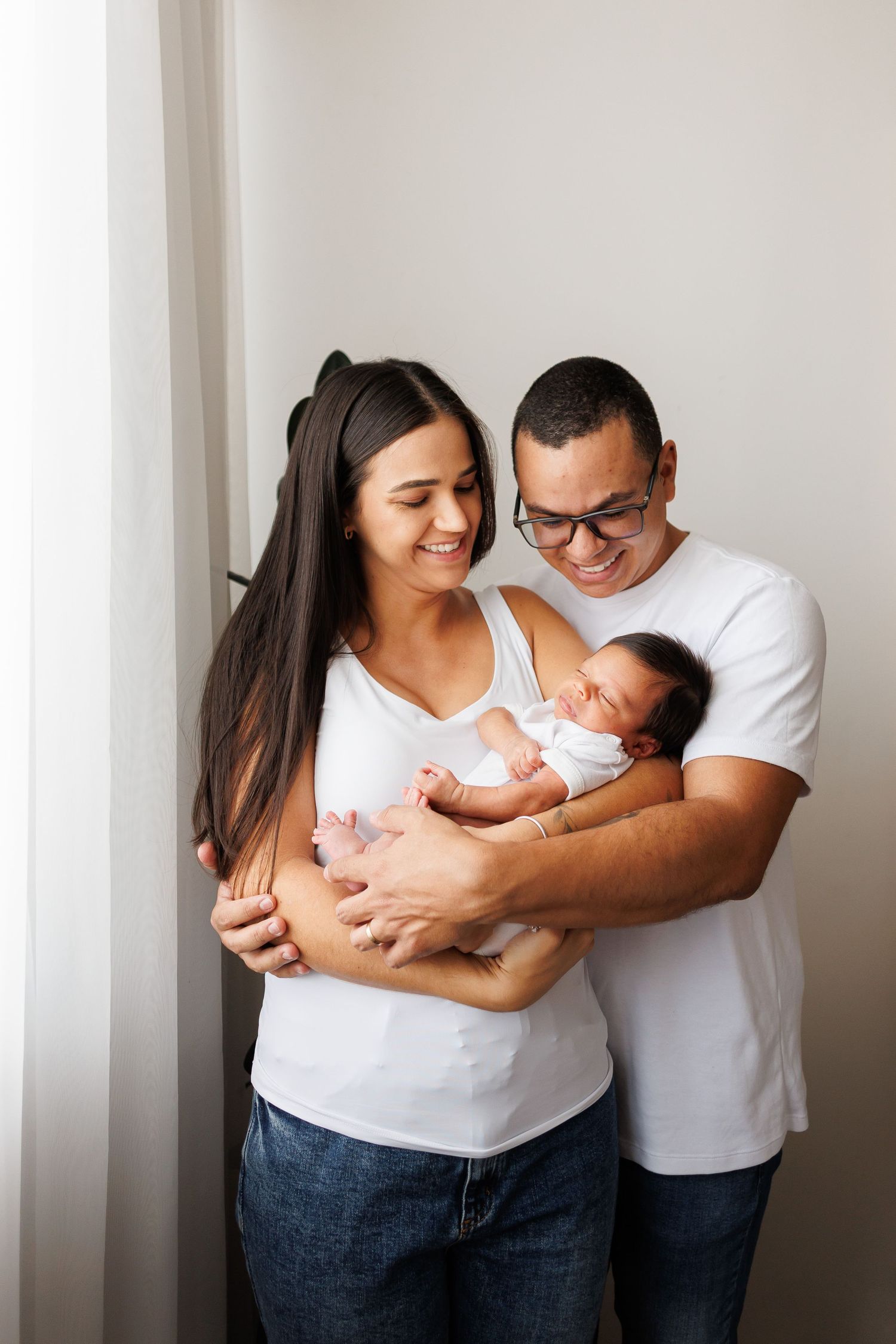 Happy young parents in white shirts smiling and holding their newborn baby together near a bright window.