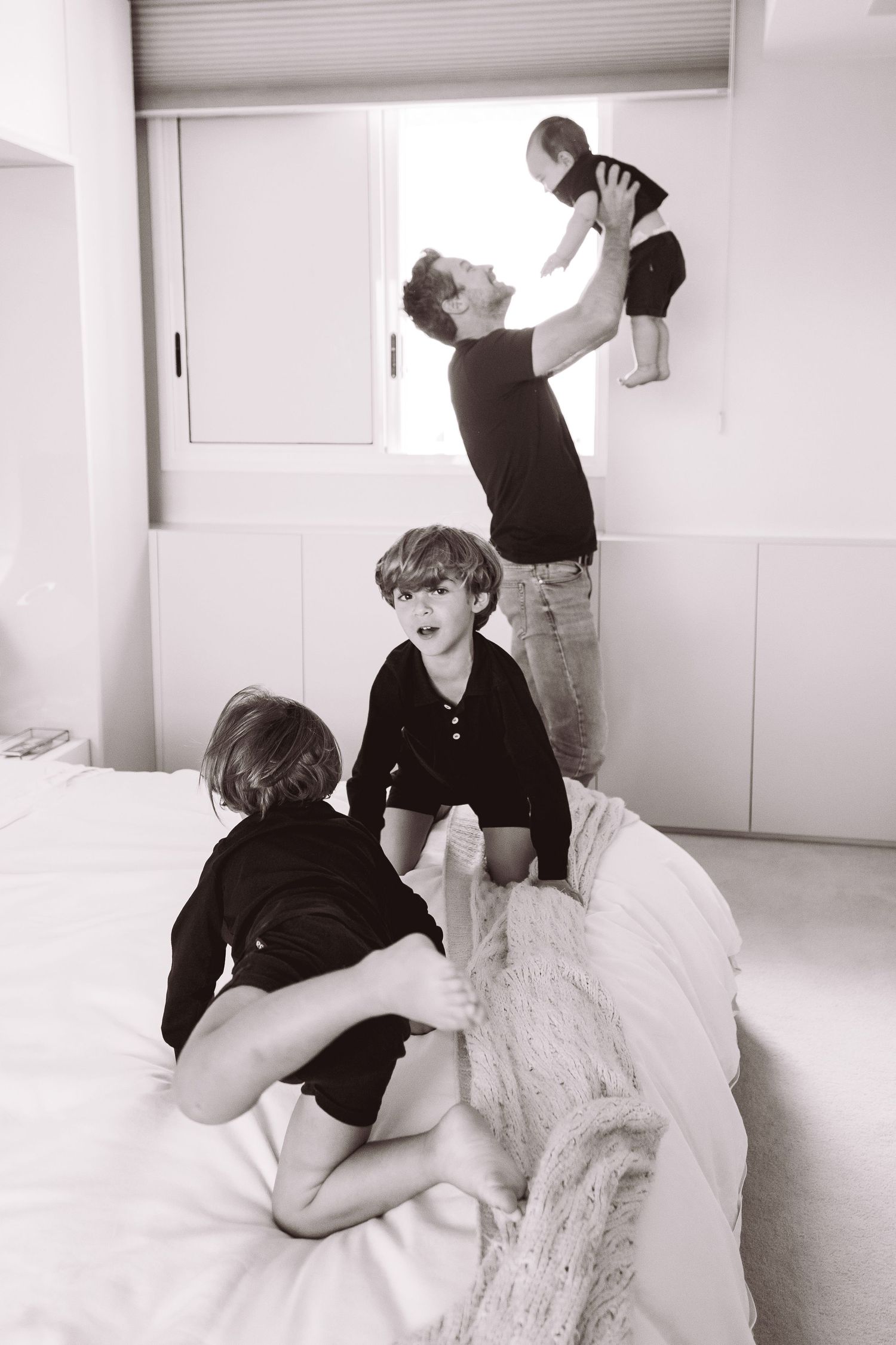 Father lifts baby near window while two children play on white bed in black and white family photo.