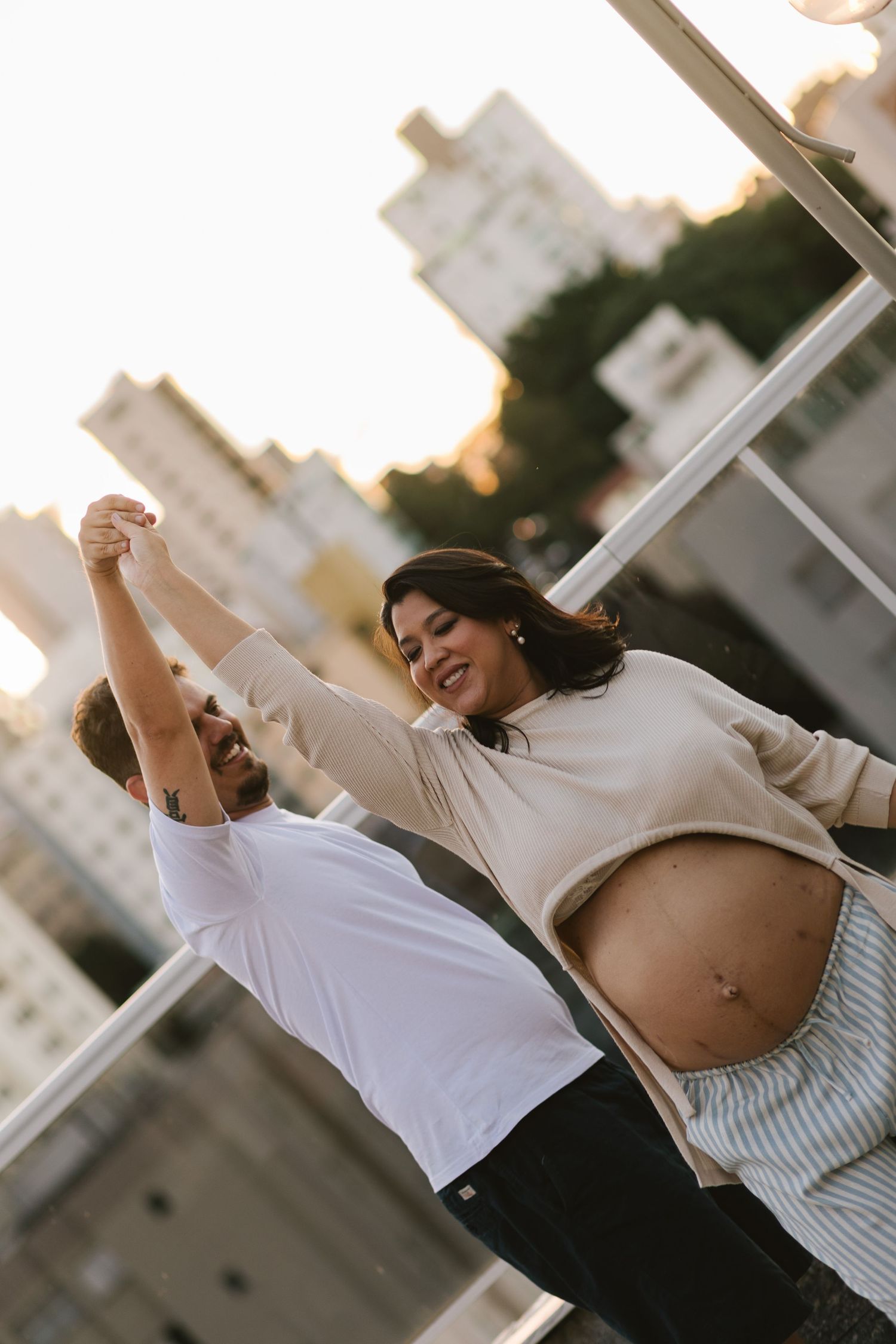 Happy pregnant couple dancing outdoors at sunset, woman showing baby bump, city buildings in background.