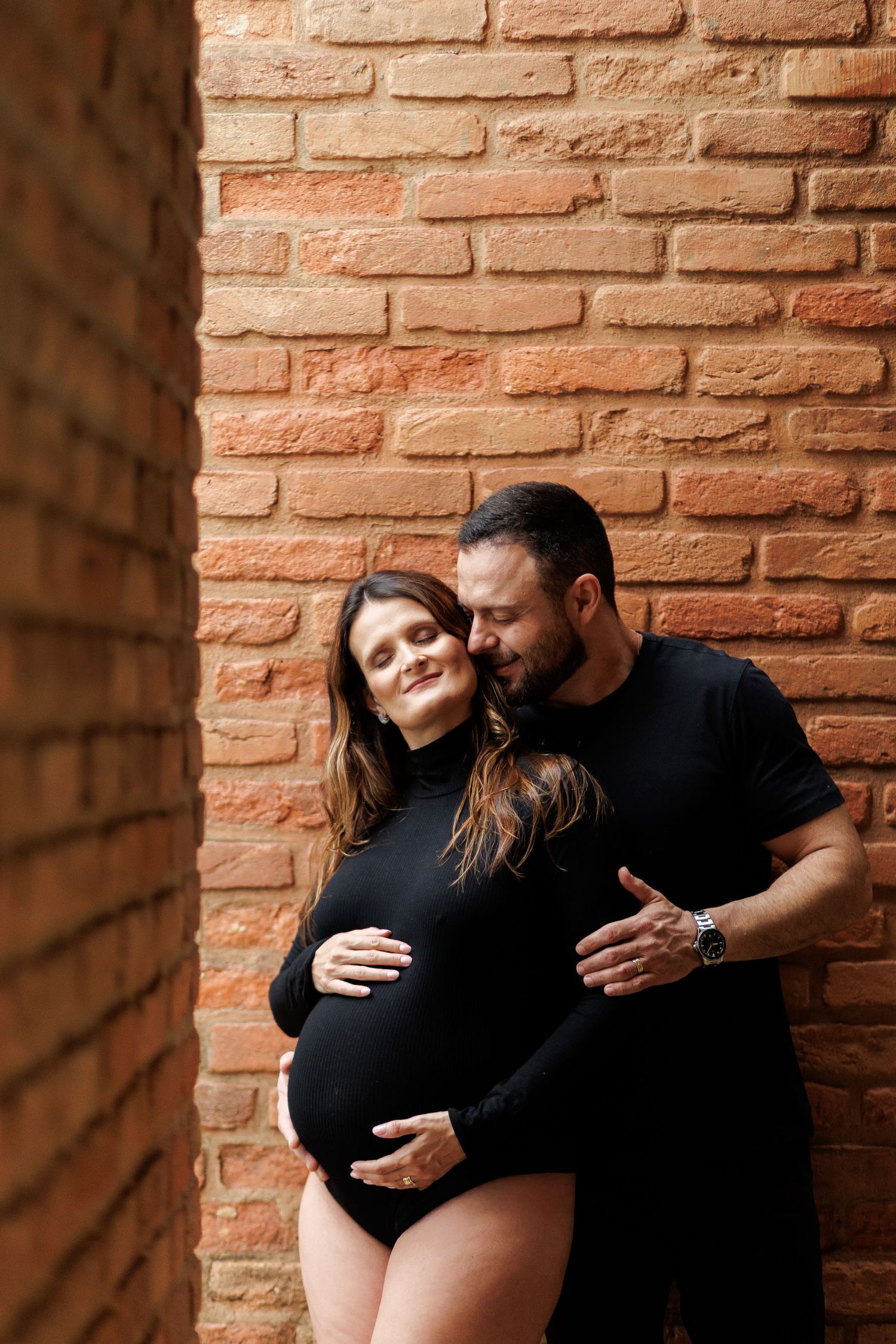 A couple in black outfits pose against a brick wall, man tenderly embracing pregnant woman from behind.