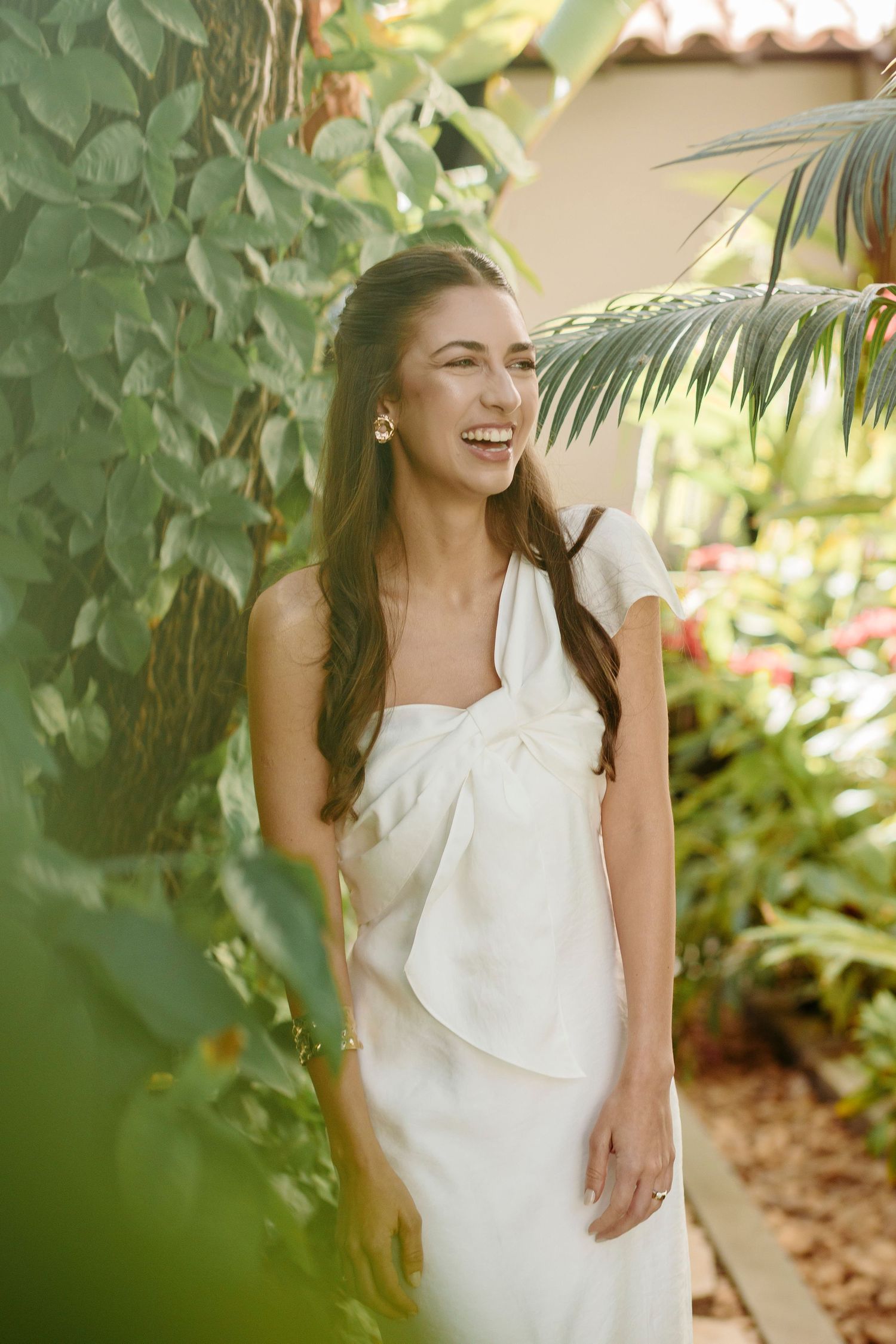 A woman in a white one-shoulder dress poses in a tropical garden setting.