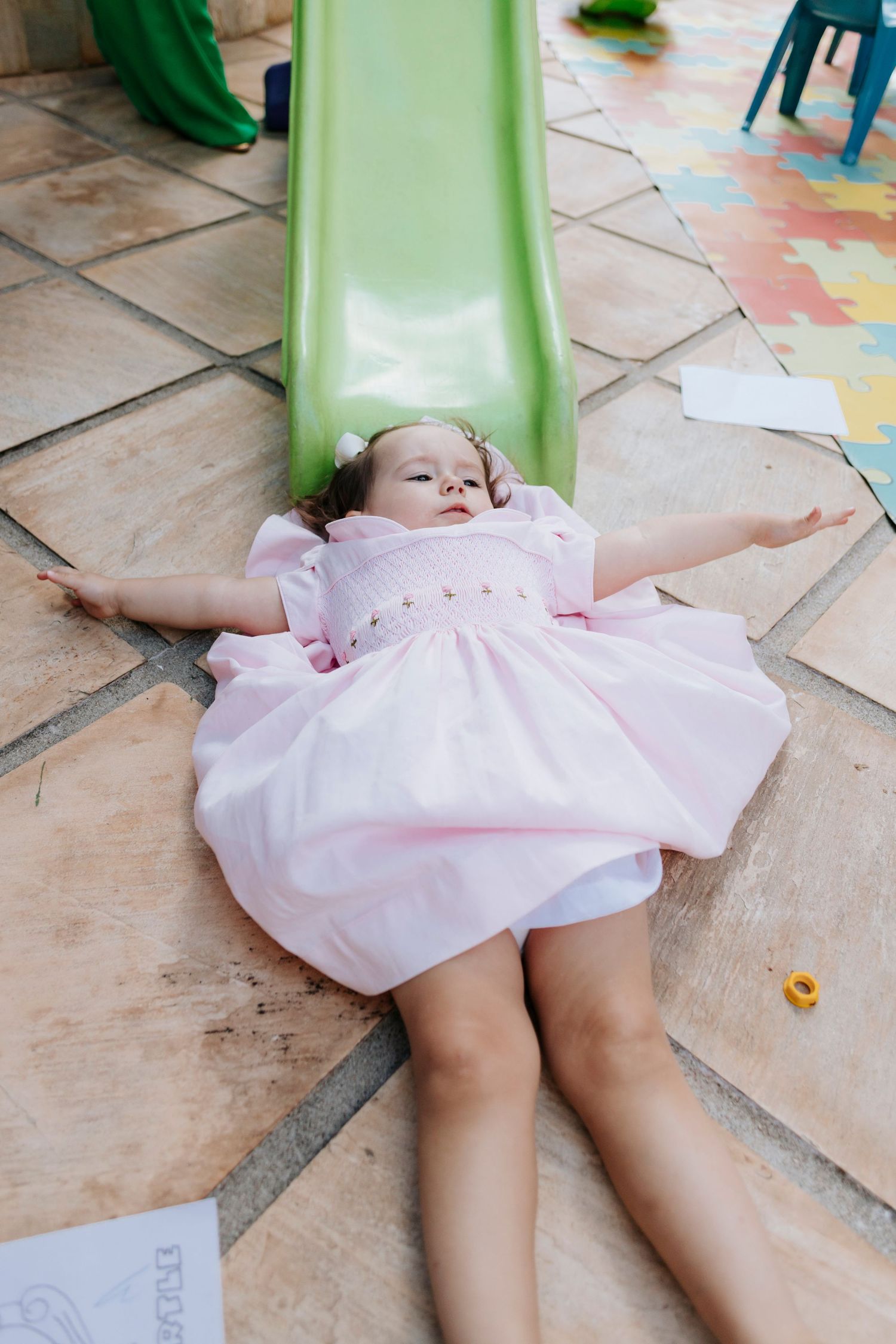 A child in a pink dress lies back on a green slide with arms spread wide.