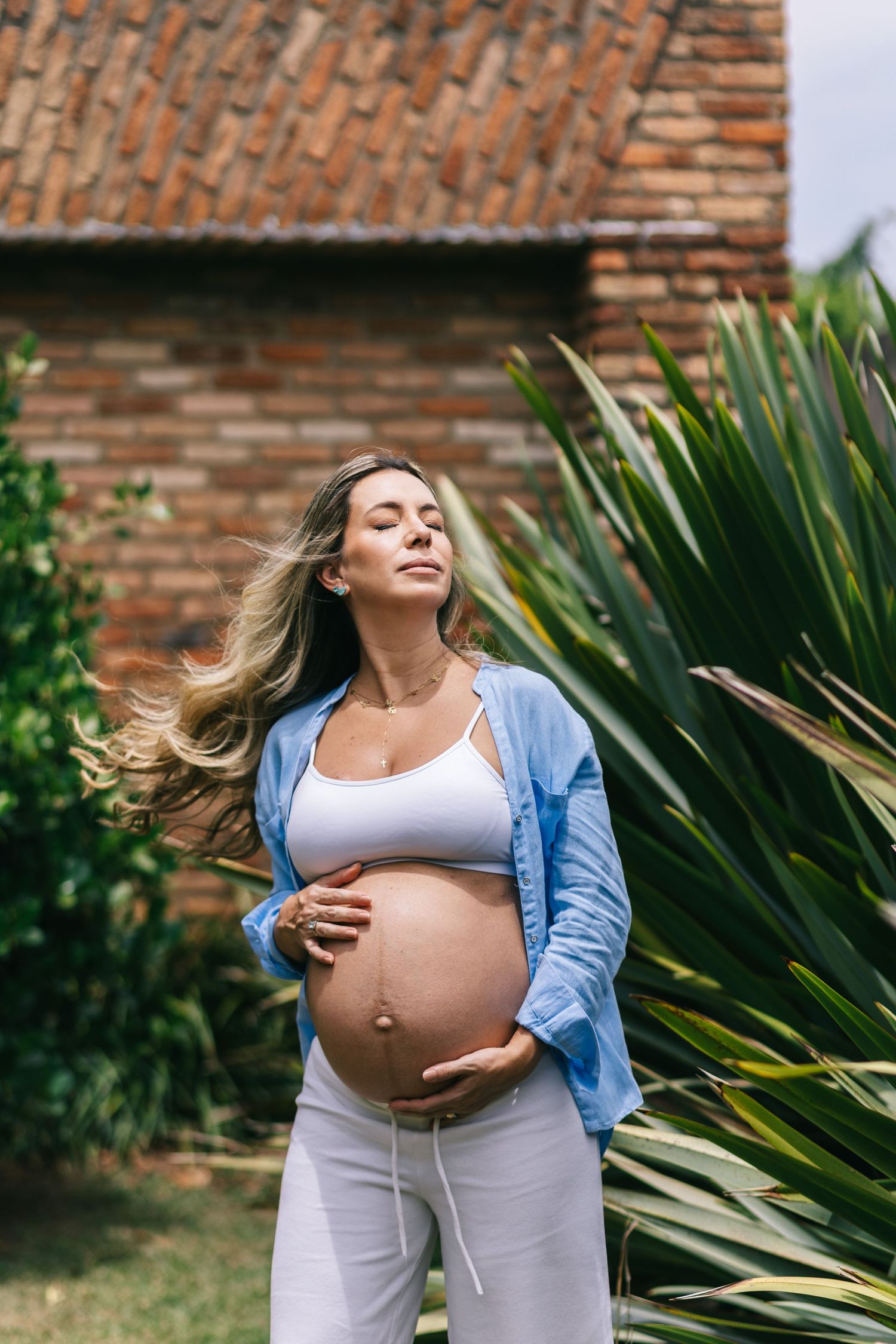 Expectant mother in denim shirt and white pants stands in garden with brick wall and tropical plants in background.
