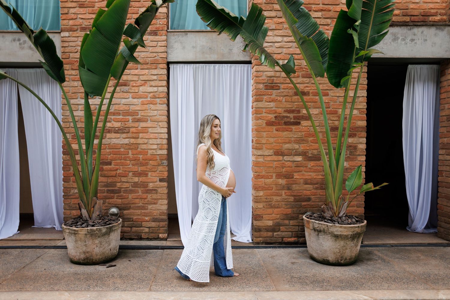 Pregnant woman in white lace dress poses between tropical plants against brick wall with white drapes.