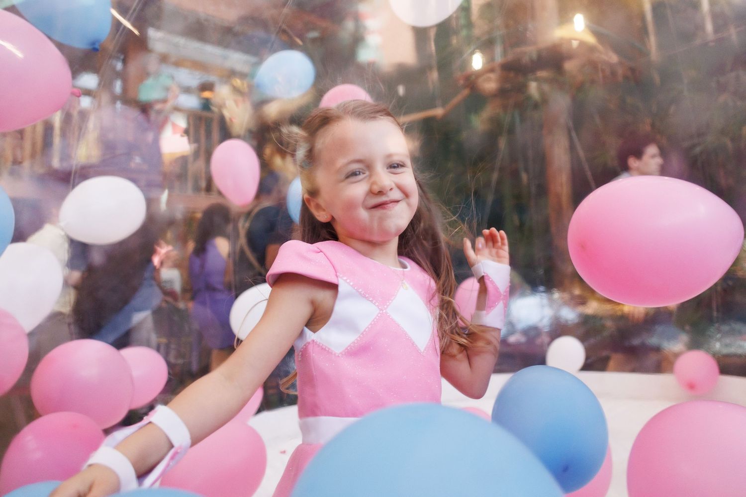 Child in pink dress celebrates at a party surrounded by pink and blue balloons in a festive atmosphere.