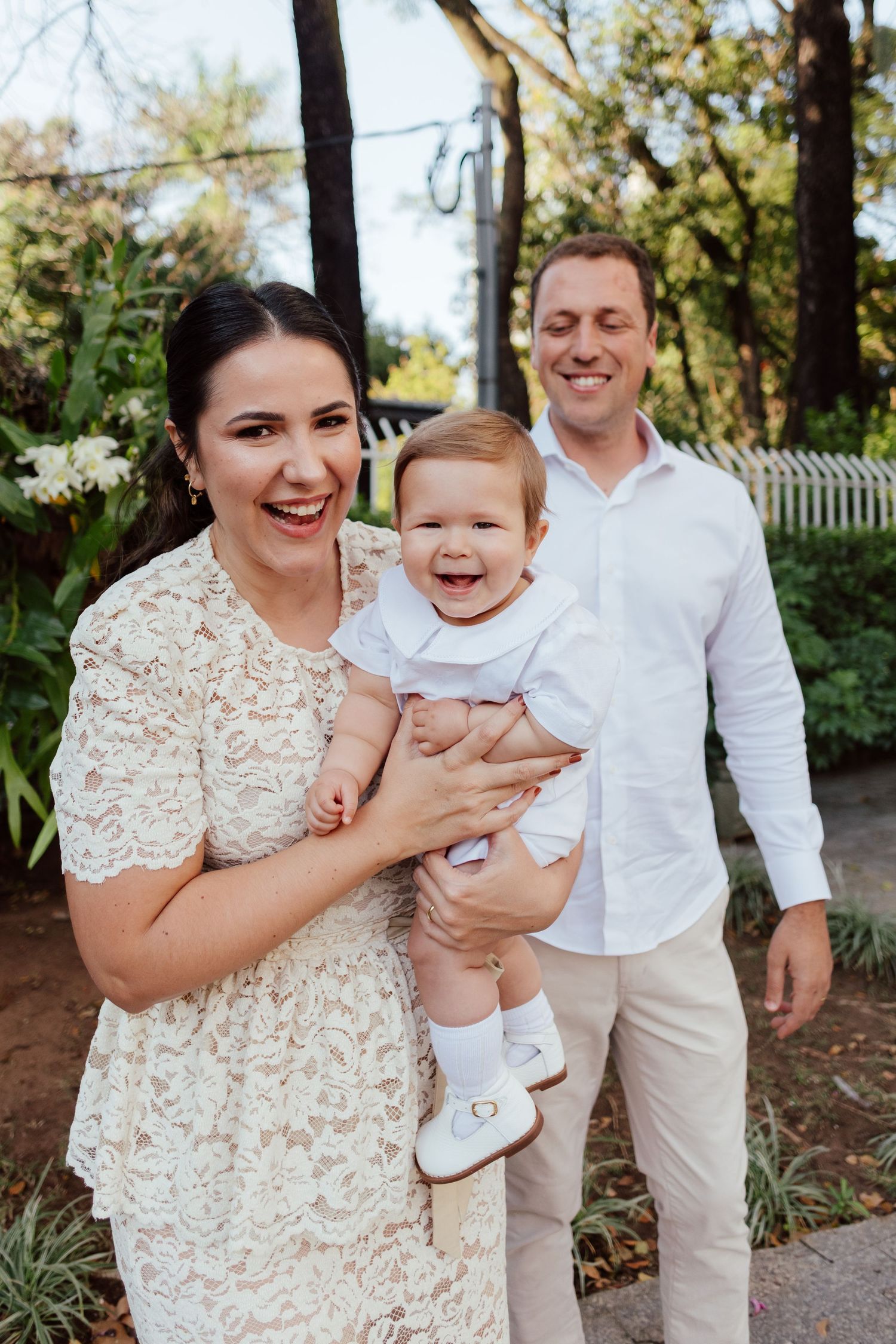 A happy family enjoying a sunny day outdoors, with a laughing baby in white clothing and parents in light-colored attire.