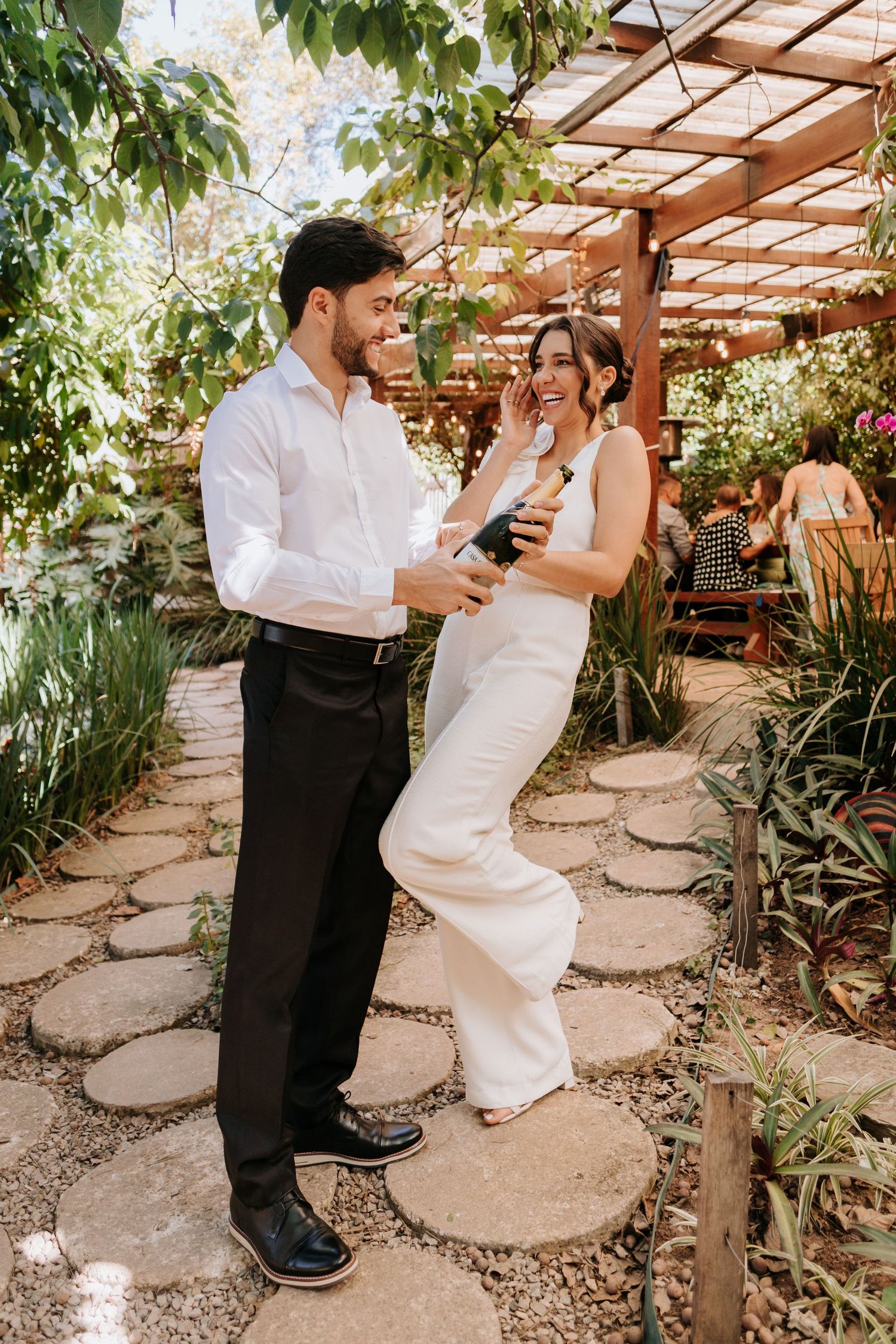 A couple in matching white outfits sharing a romantic moment under a wooden pergola in a lush garden setting.