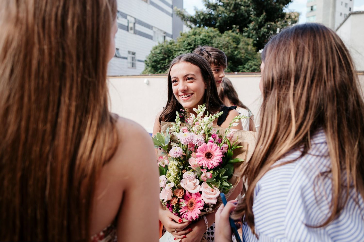 A group of friends share joyful moments while holding pink and white floral bouquets outdoors.
