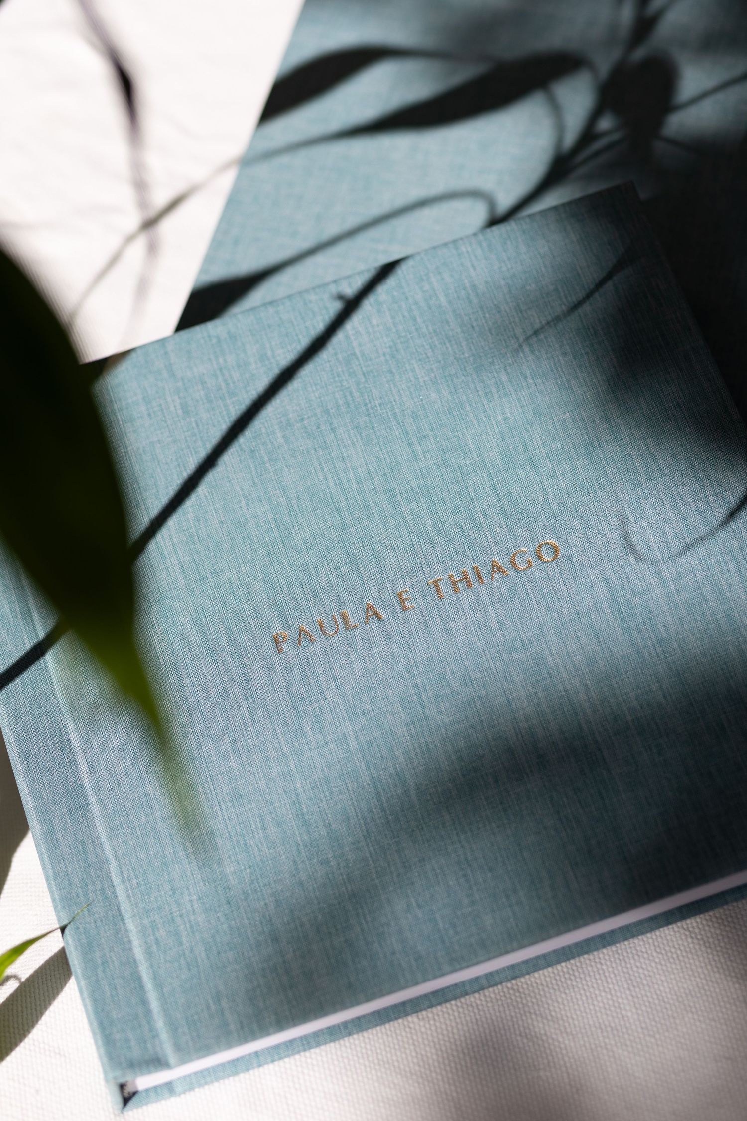 Close-up view of a grey hardcover wedding album with gold text showing 'Paula e Thiago' on the cover.