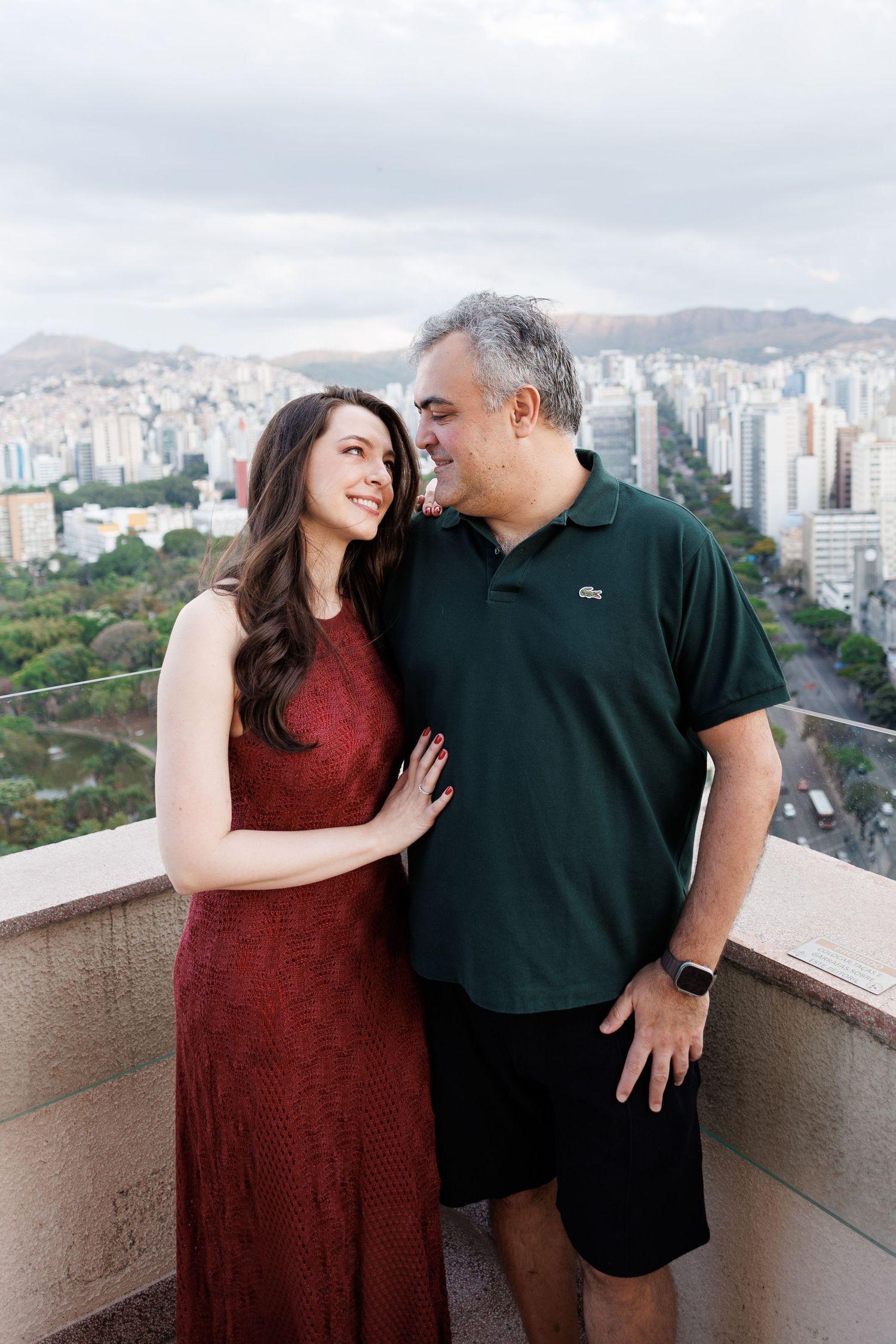 Two people share a tender moment on a rooftop balcony overlooking a cityscape with tall buildings and mountains.