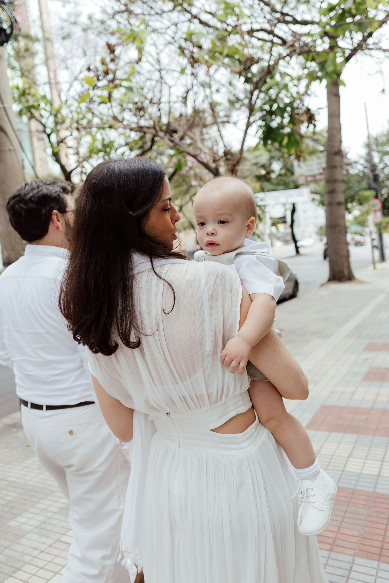 Family members in white outfits walk together on a tree-lined brick pathway on a sunny day.