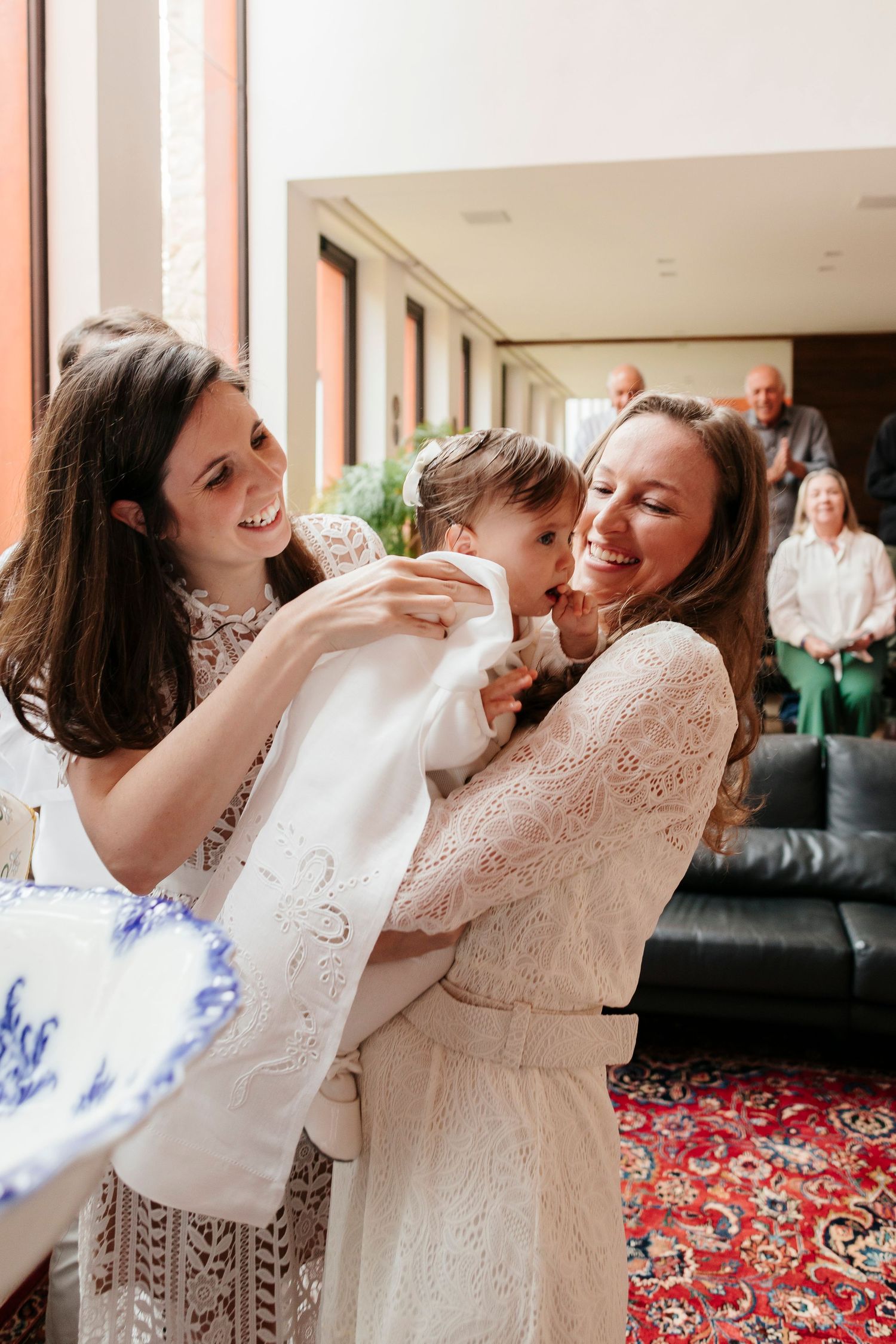 Two people in white lace dresses share a joyous moment with a baby during a christening ceremony.
