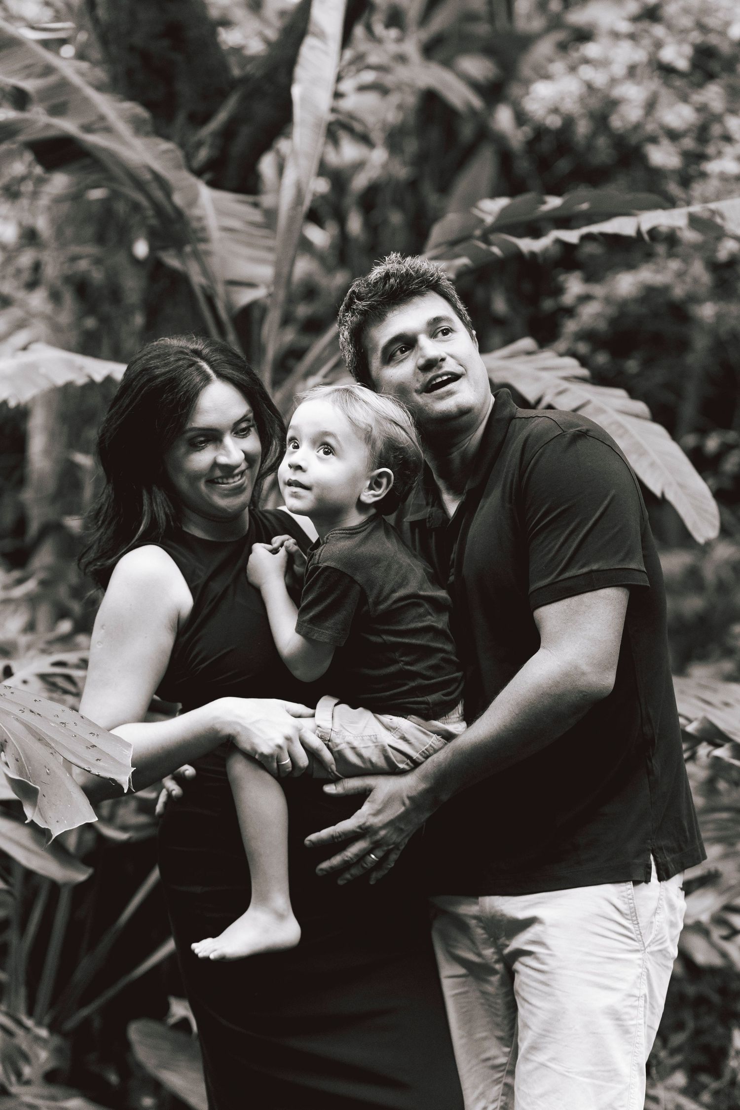 Family of three poses together in a tropical garden setting, captured in black and white.