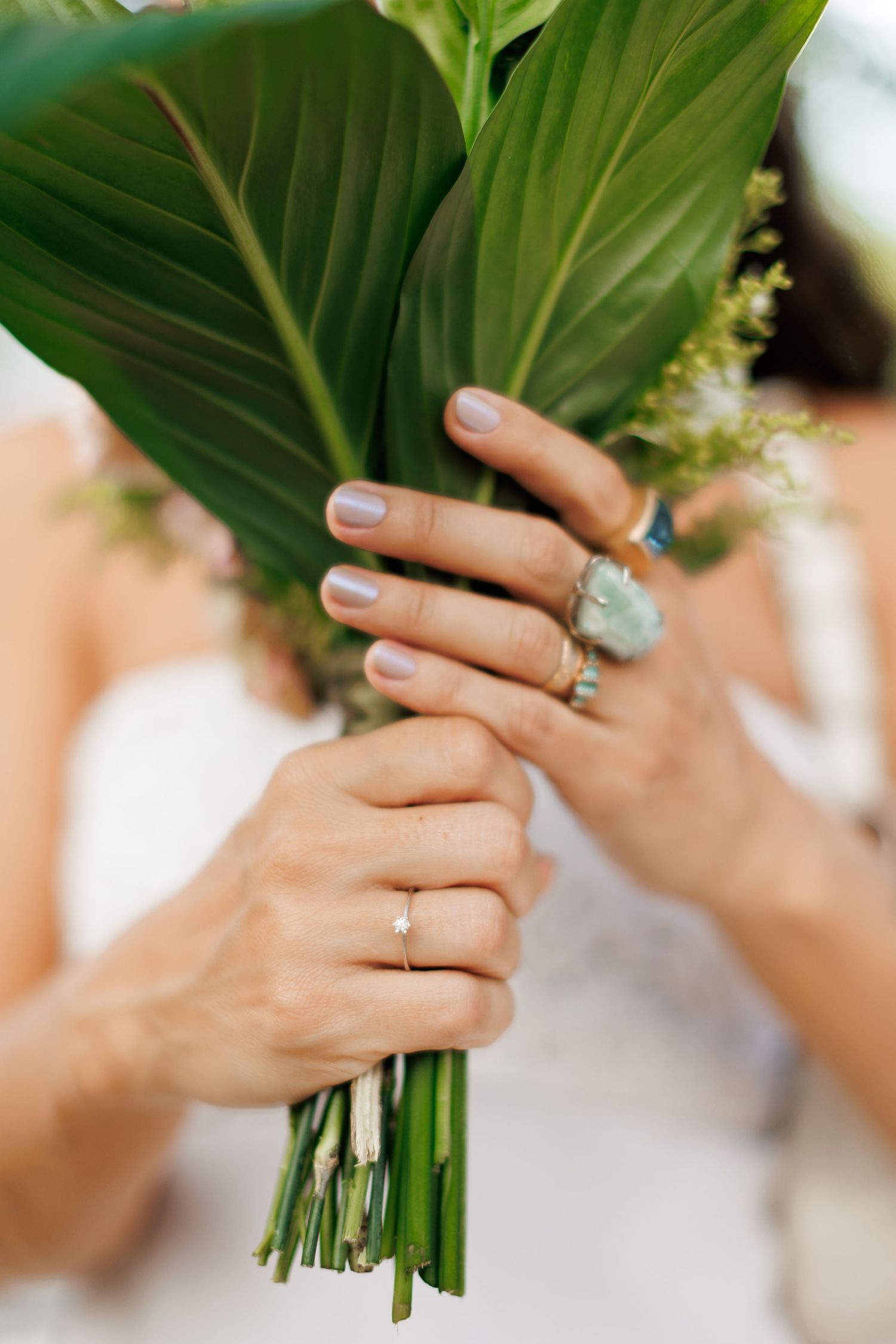 Bride holding green tropical leaf bouquet with diamond engagement ring and turquoise stone ring on fingers.