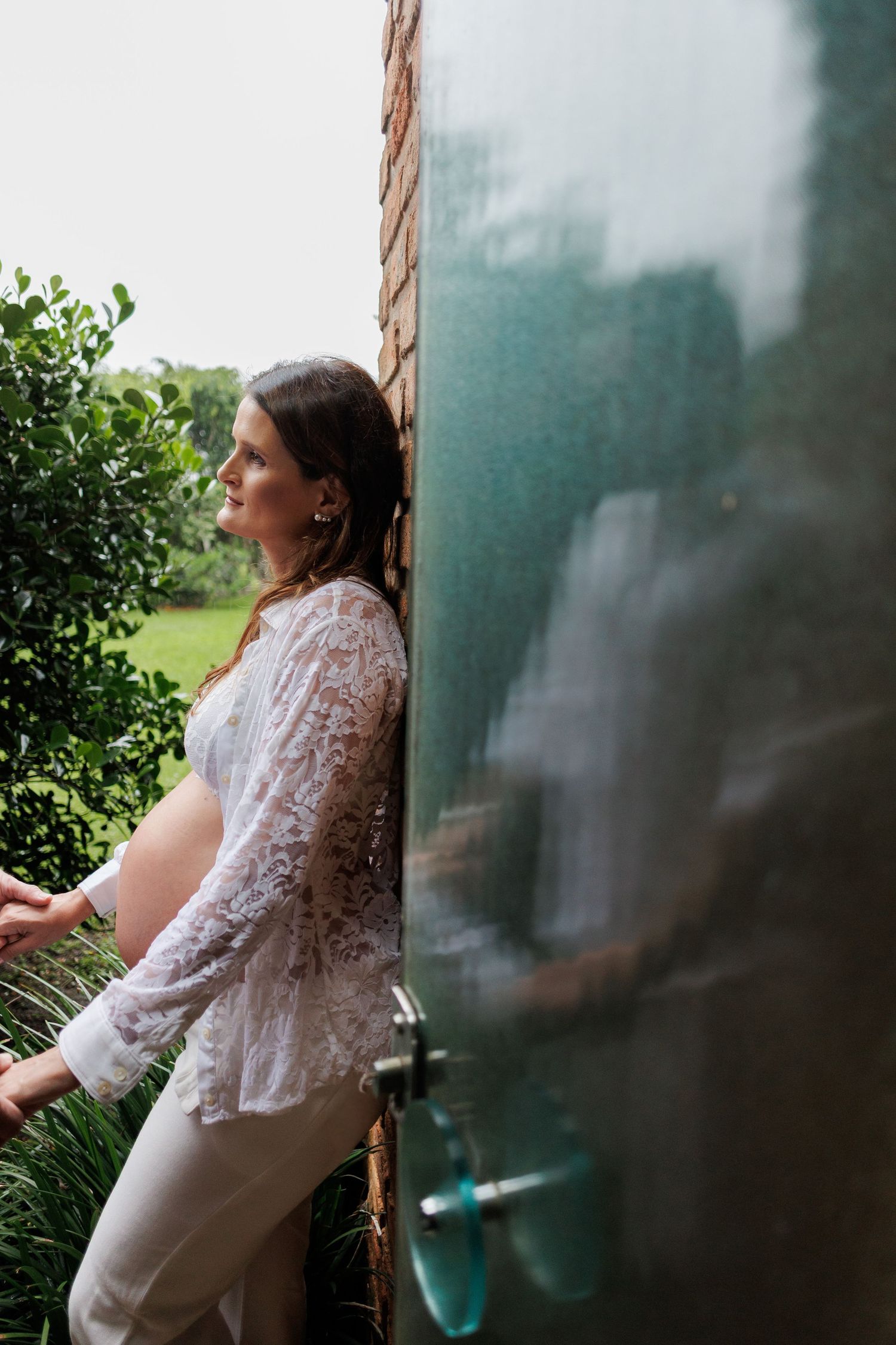 Pregnant woman in white lace top leaning against brick wall beside glass door in garden setting.