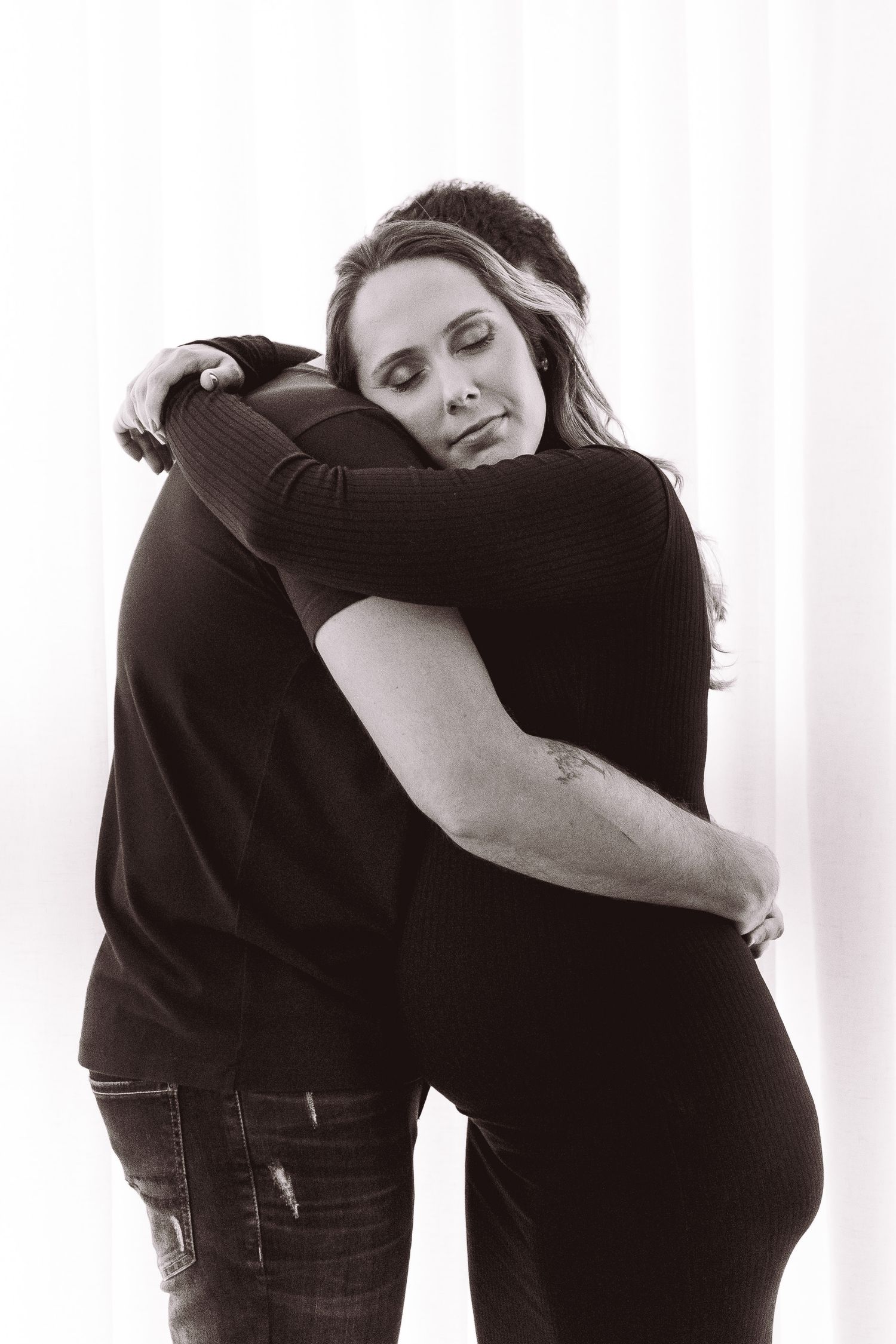 Black and white photo of two people sharing a warm, emotional embrace against a soft white curtain background.