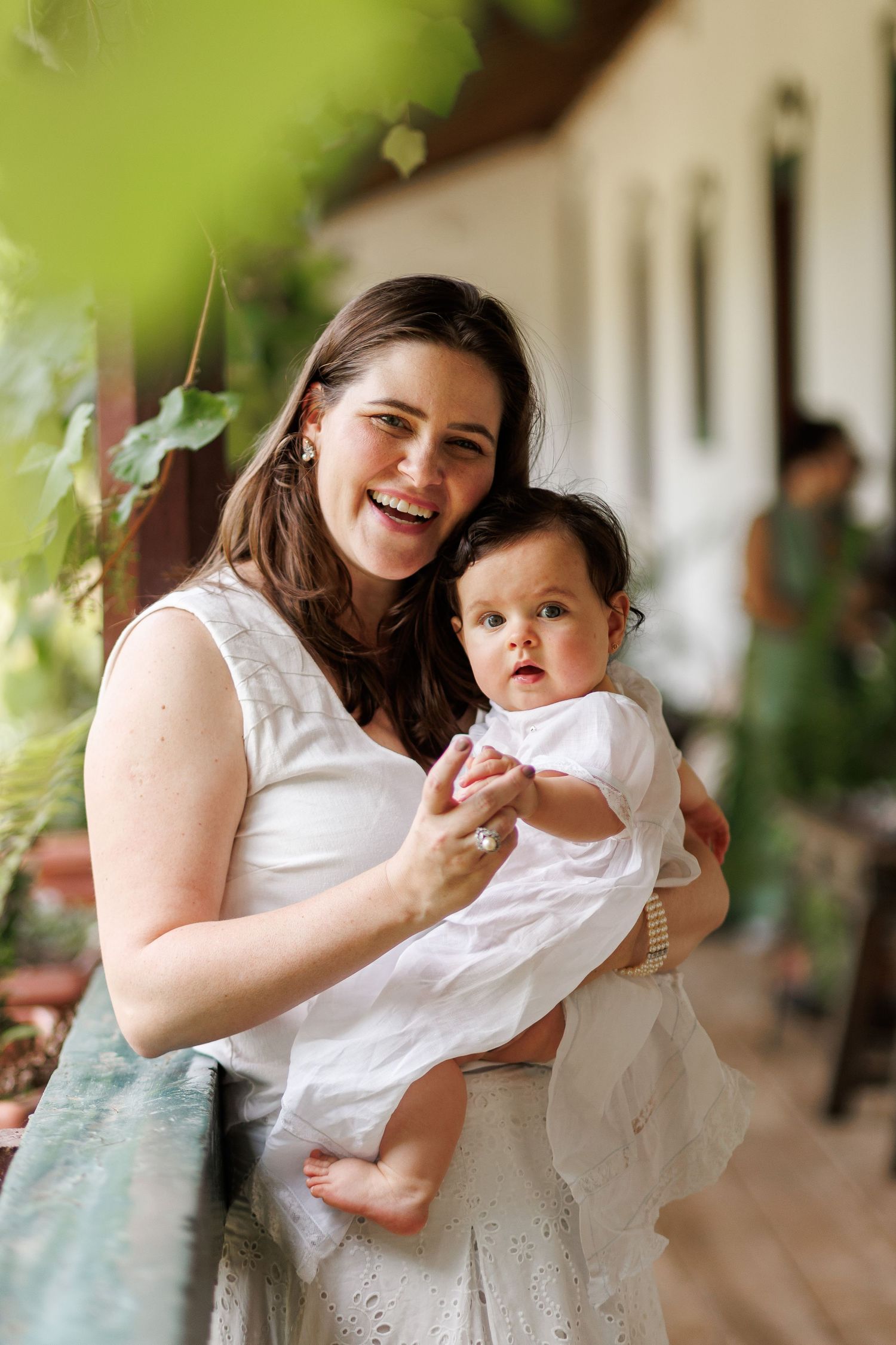 Smiling woman in white dress holding adorable baby in white outfit on a sunlit porch.