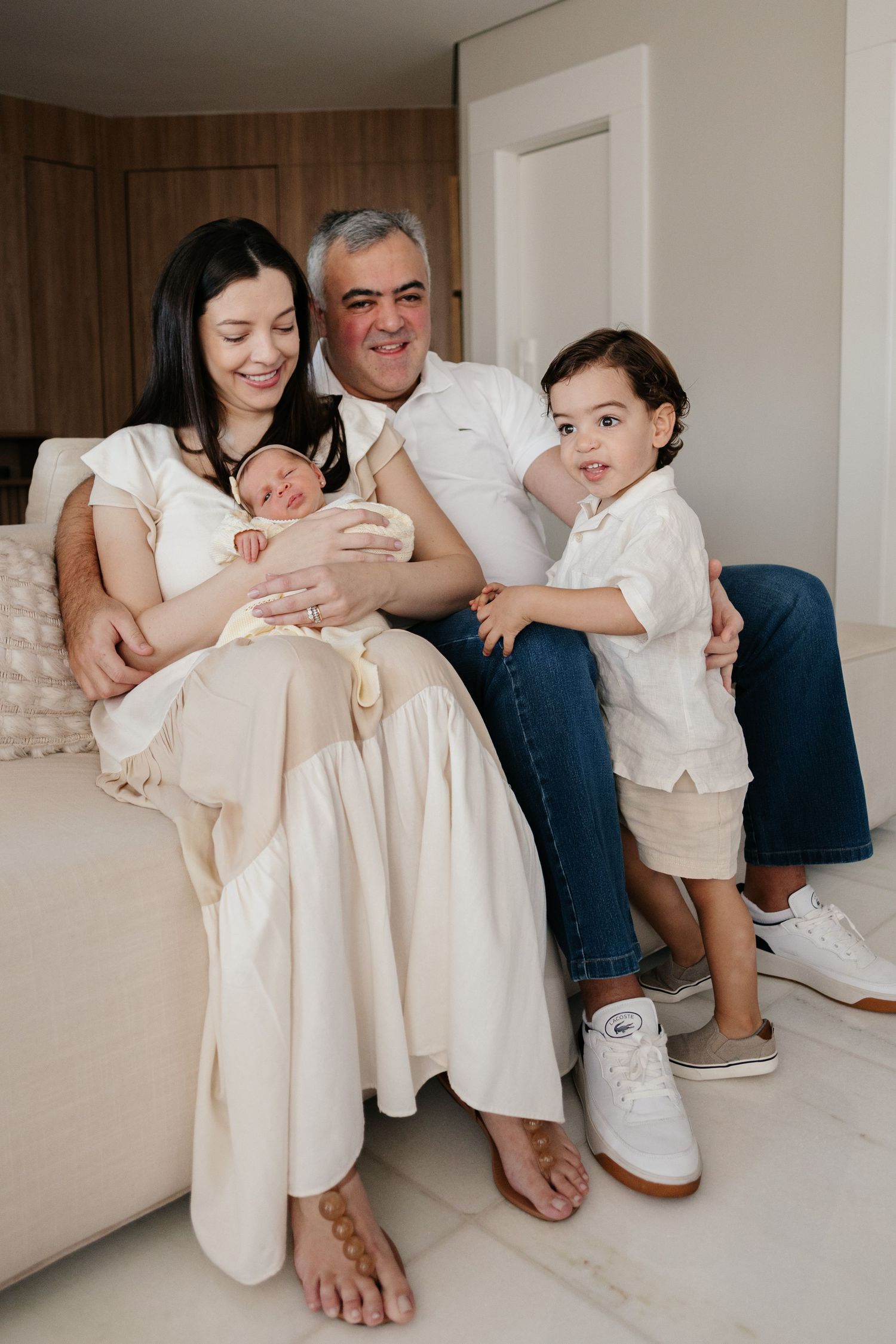 Happy family portrait with parents, newborn baby, and toddler sitting together on a couch indoors.