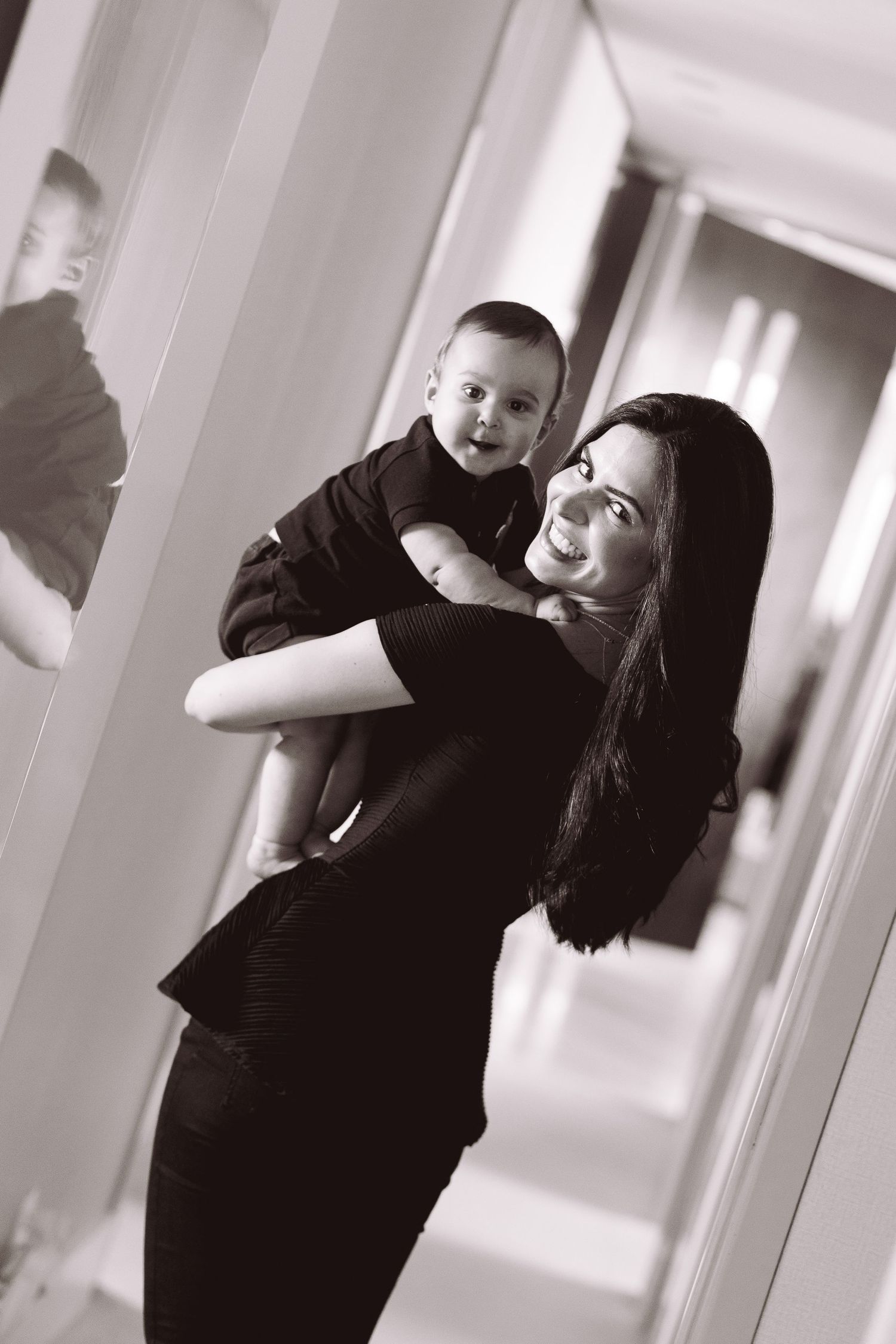 Mother smiles holding happy baby in hallway, captured in elegant black and white portrait photo.