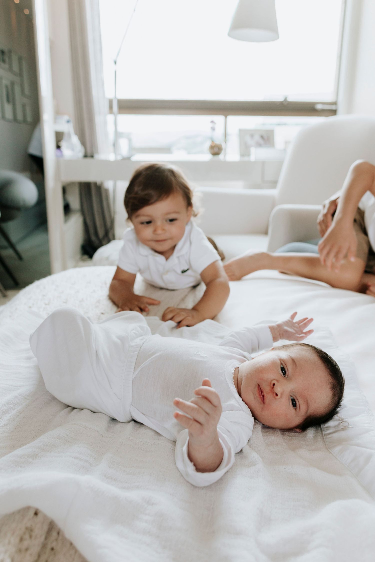 Two siblings in white outfits playing together on bed in bright room.