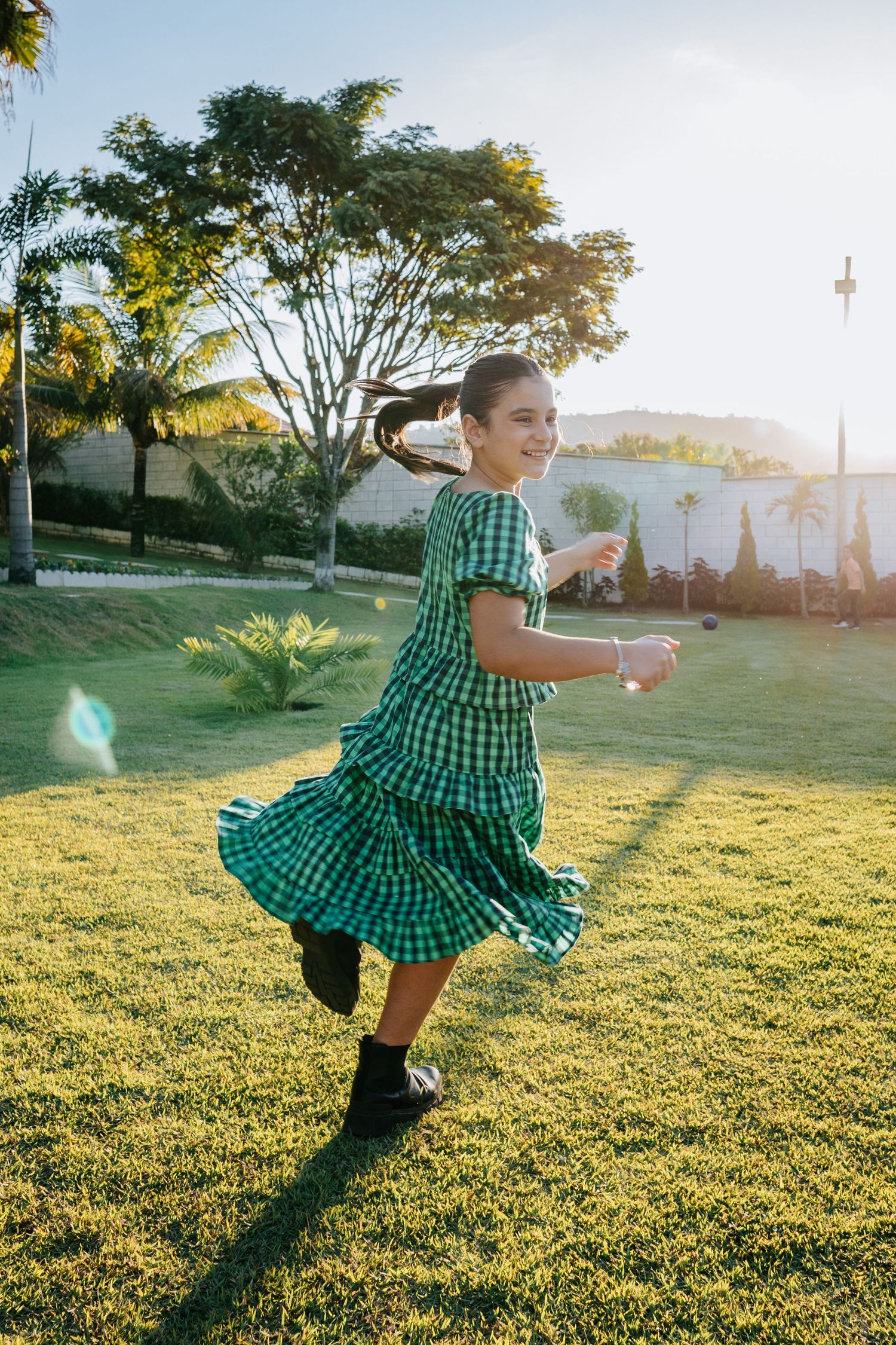 A person in a green gingham dress twirls joyfully on a sunlit lawn with trees in the background.