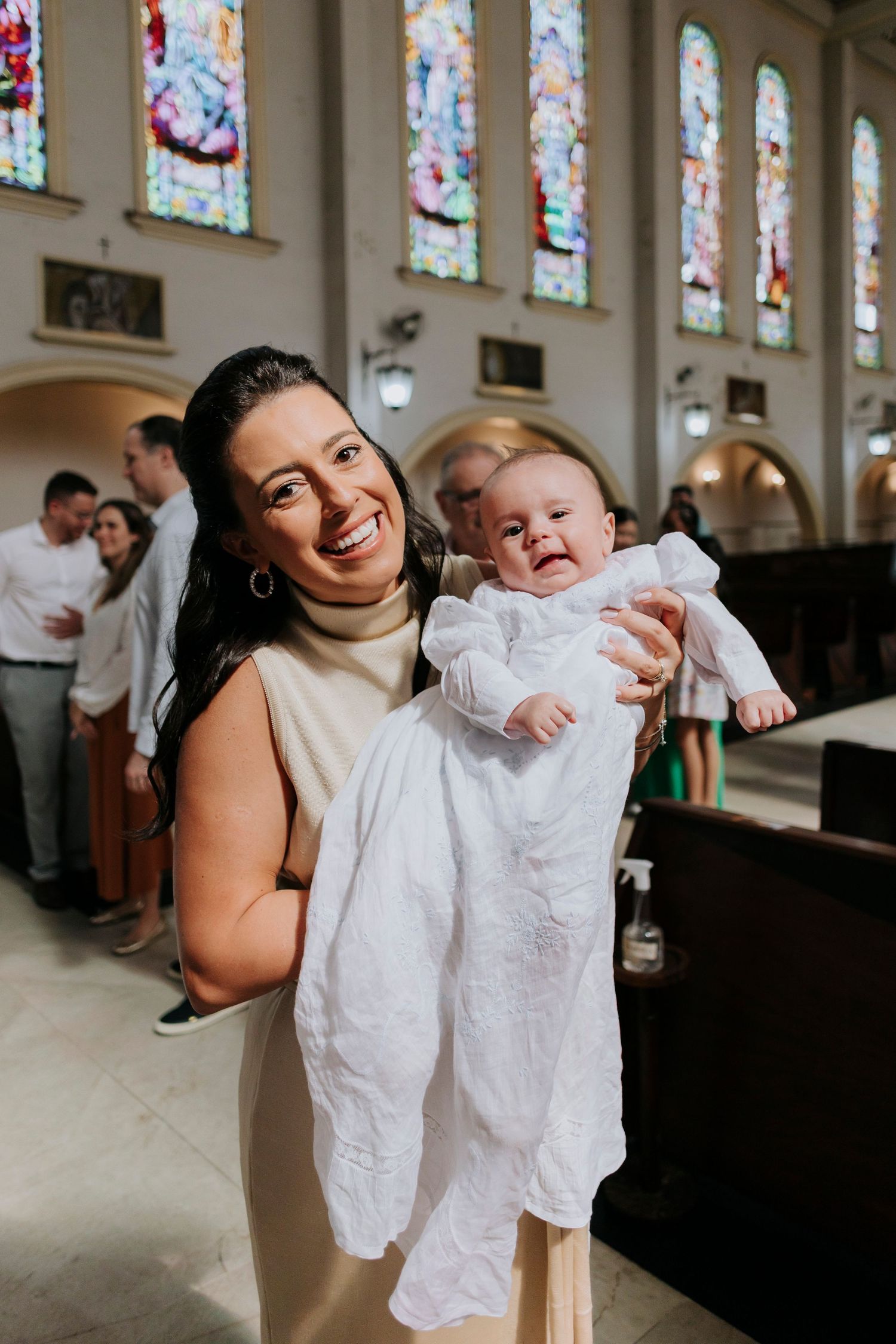 A joyful moment inside a church with stained glass windows as a baby in white christening attire is held during the ceremony.