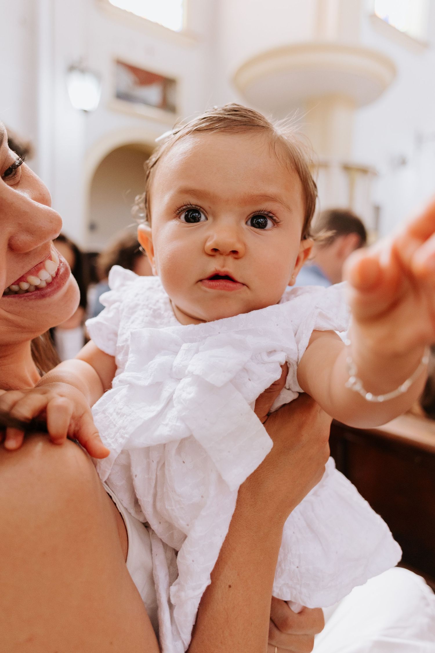Baby in white dress being held during a special celebration in a bright indoor setting.